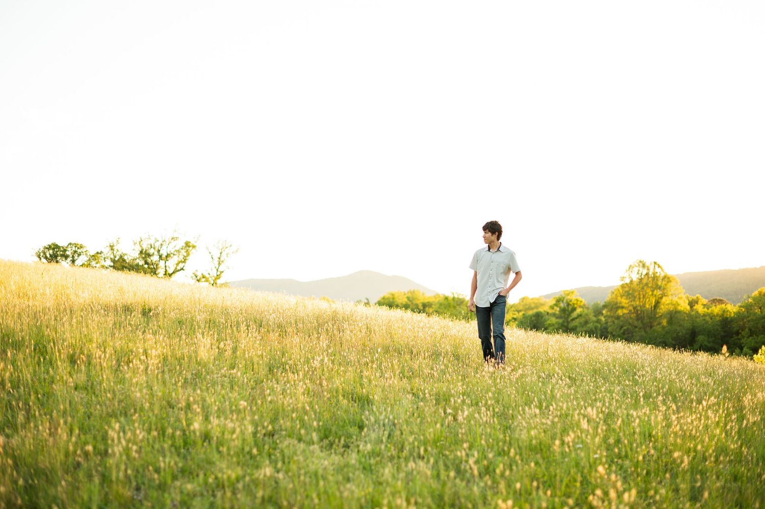 A man walks through a lush green meadow with golden grass under bright sunlight with rolling hills in the background.