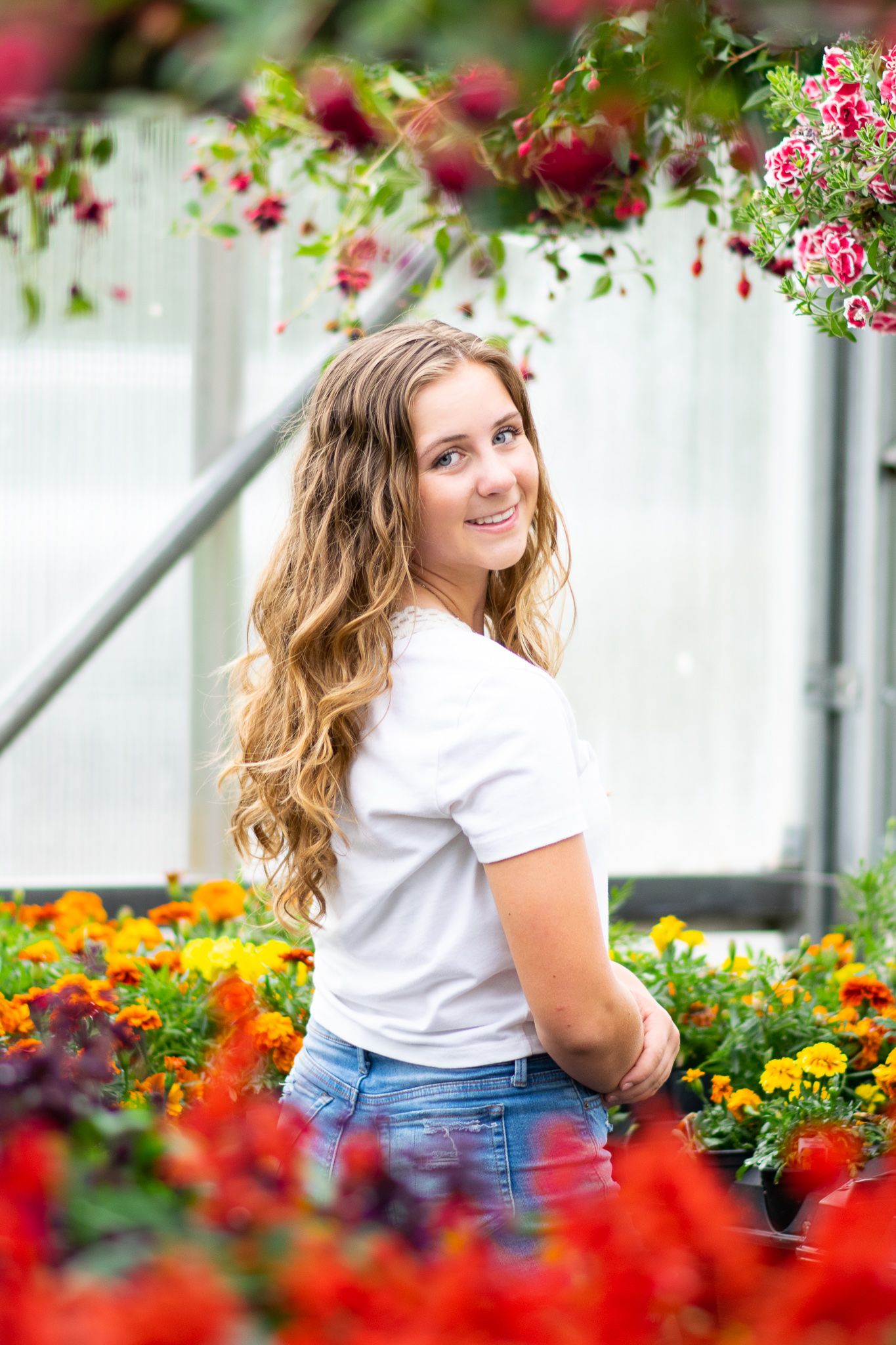Young woman with long curly blonde hair smiling in a colorful greenhouse surrounded by vibrant red, orange, and yellow flowers.
