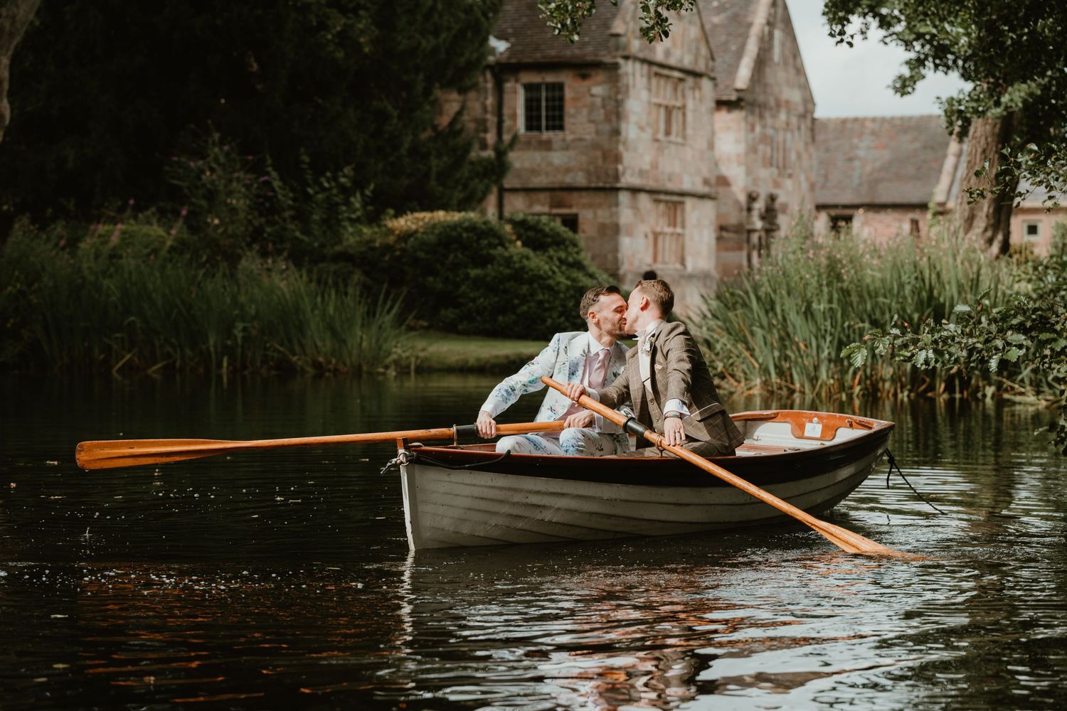 Couple sharing a romantic moment in a wooden rowboat on a calm lake beside a historic stone manor.