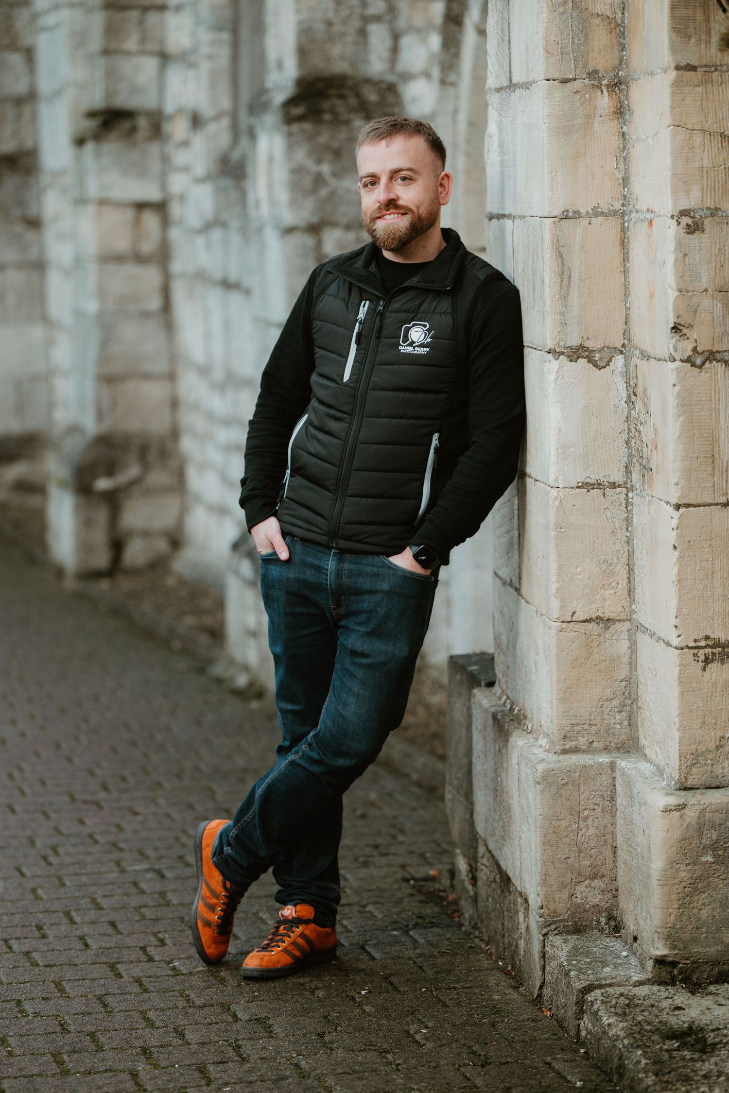 A person in a black puffer jacket and jeans leans against a stone wall while wearing bright orange sneakers.