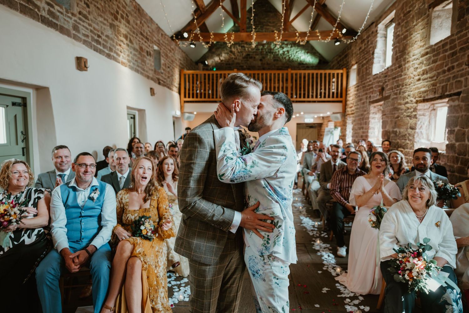 Wedding guests celebrate and dance together inside a rustic barn venue with exposed brick walls and wooden beams.