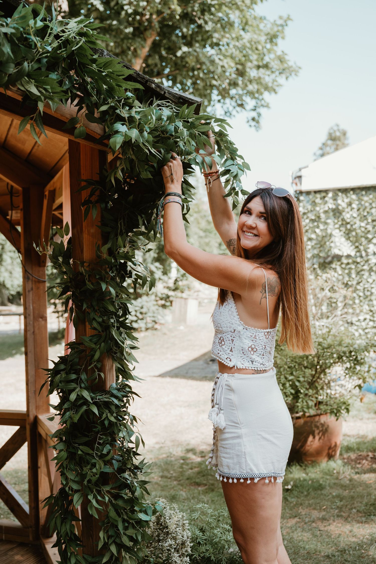 A person in a white lace crop top and shorts decorates a wooden pergola with fresh greenery on a sunny summer day.