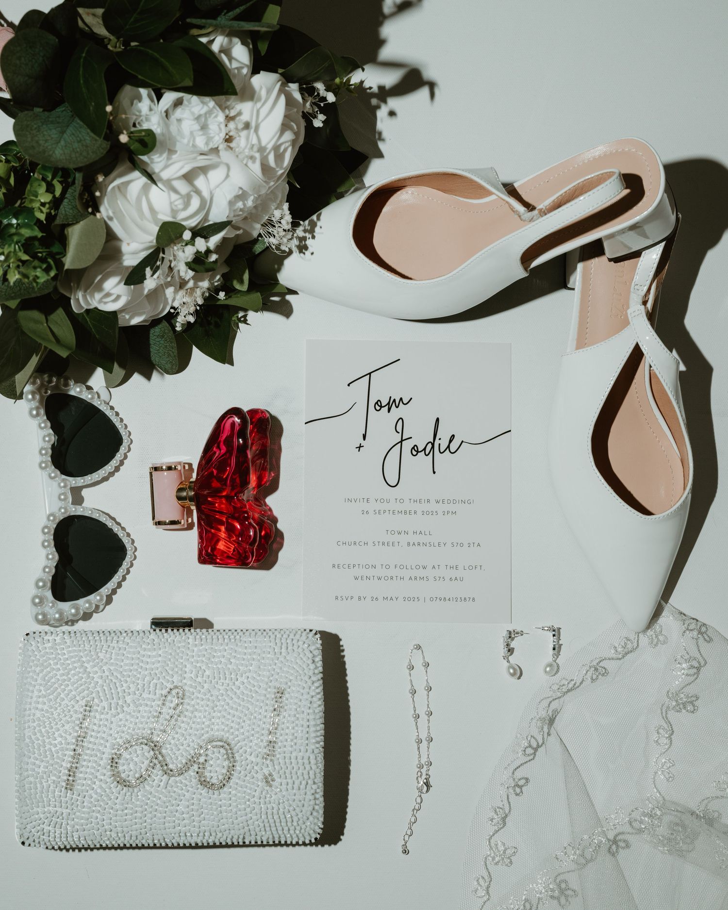 Wedding accessories including a beaded clutch, invitation cards, and floral decorations arranged on a white surface.