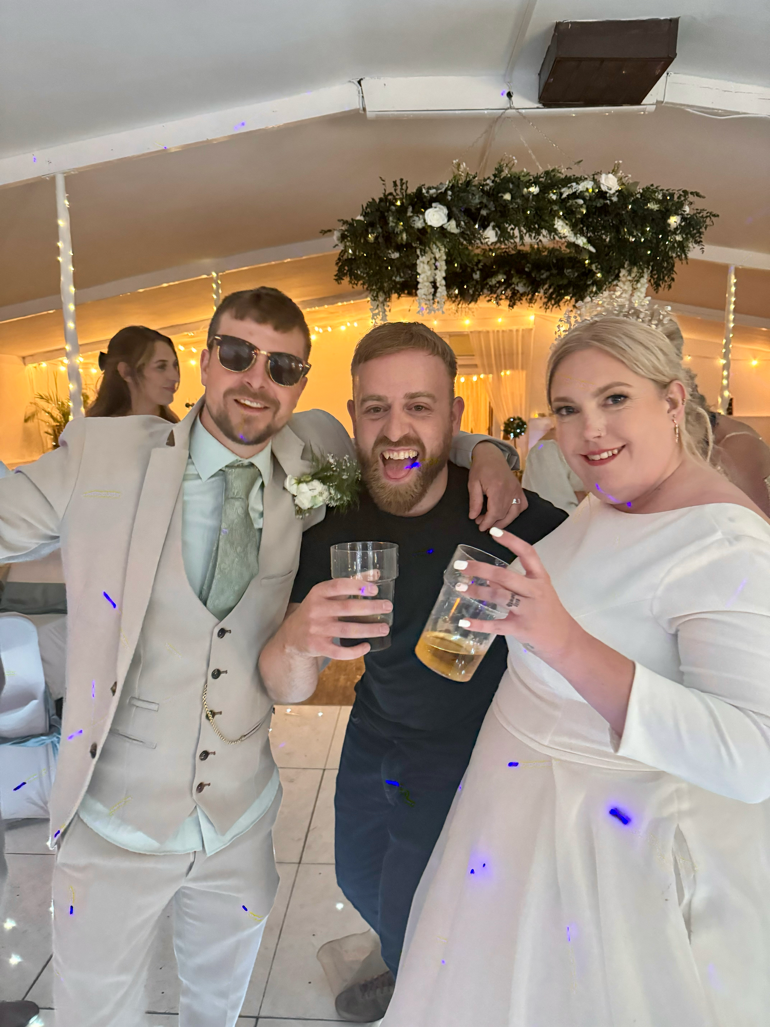 A group celebrates at a wedding reception with drinks and laughter in a white-decorated venue with greenery accents.