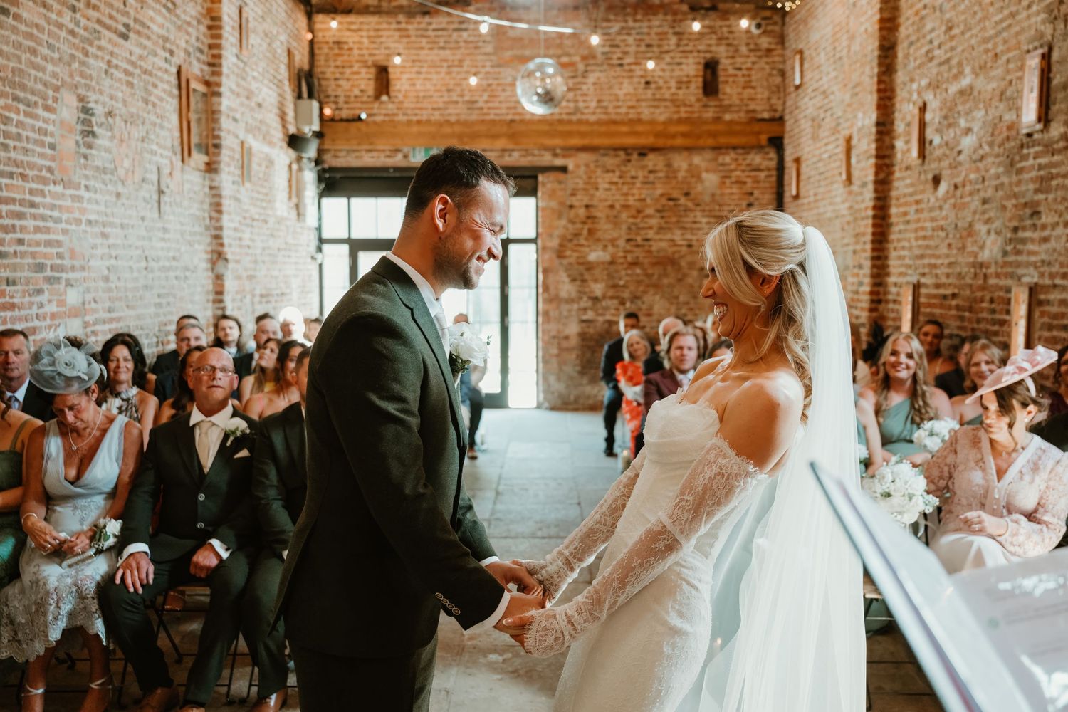 Bride and groom holding hands at rustic brick wedding venue ceremony with guests seated in background.