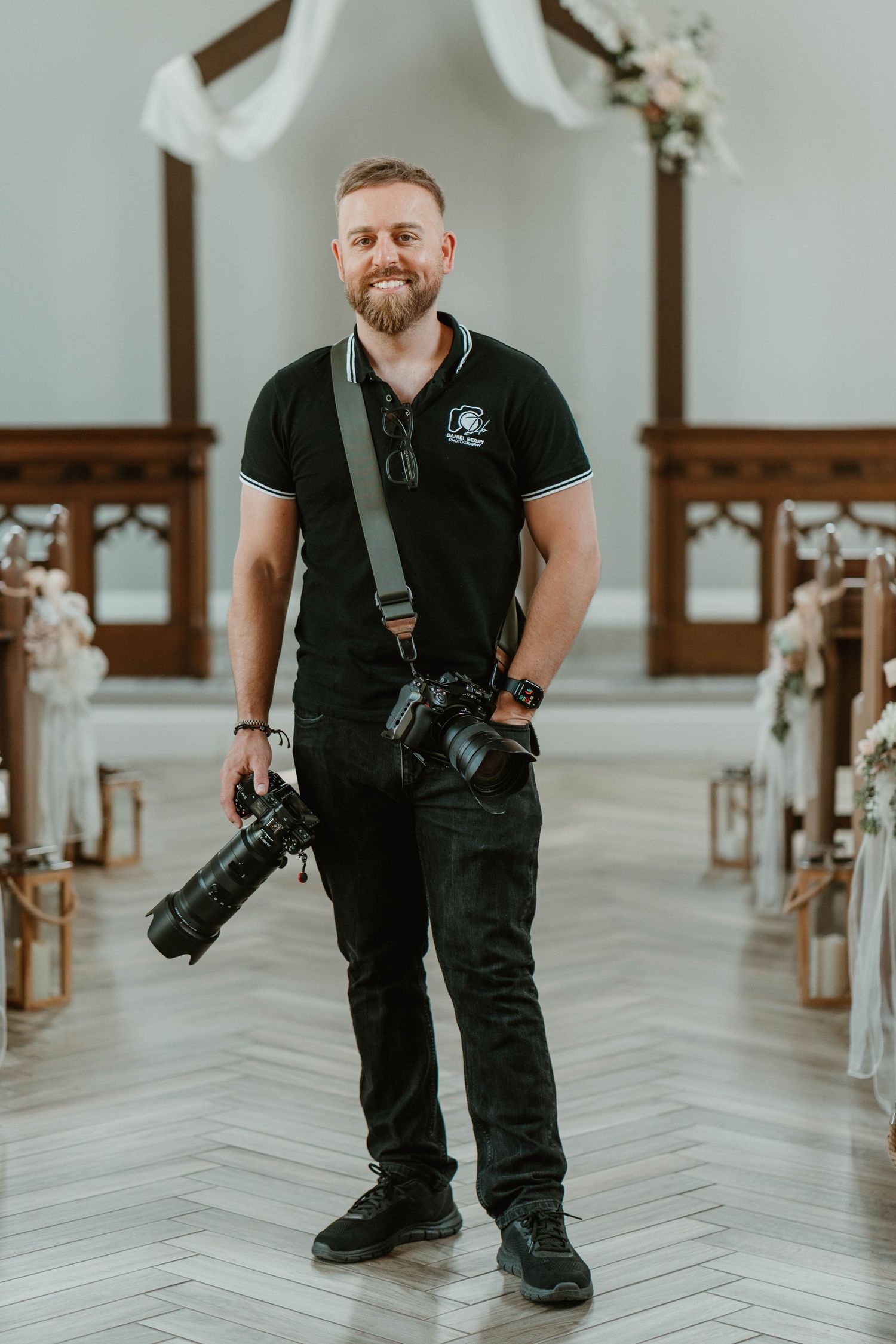 Smiling male wedding photographer in black polo holding cameras inside an elegant chapel with floral decorations.