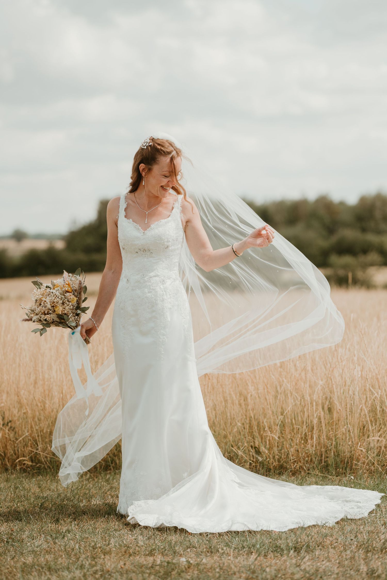 Bride in white lace gown holds bouquet and veil in golden field on a cloudy day.