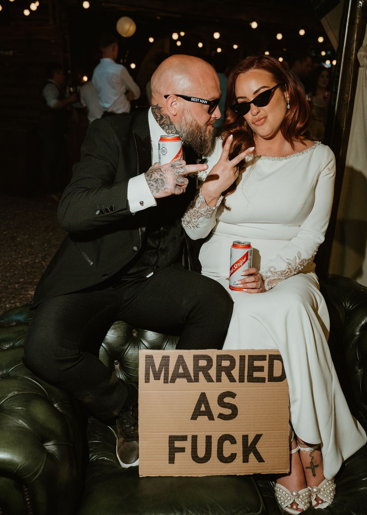 Tattooed groom and bride in white dress holding beer cans, sitting with a cardboard sign reading Married As Fuck.