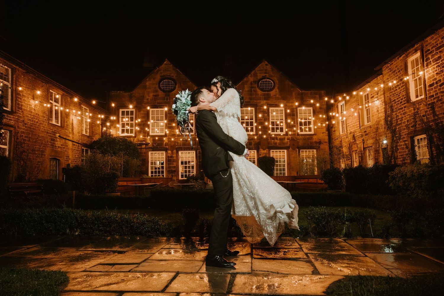 A couple dances under romantic string lights in a courtyard during an evening wedding celebration.