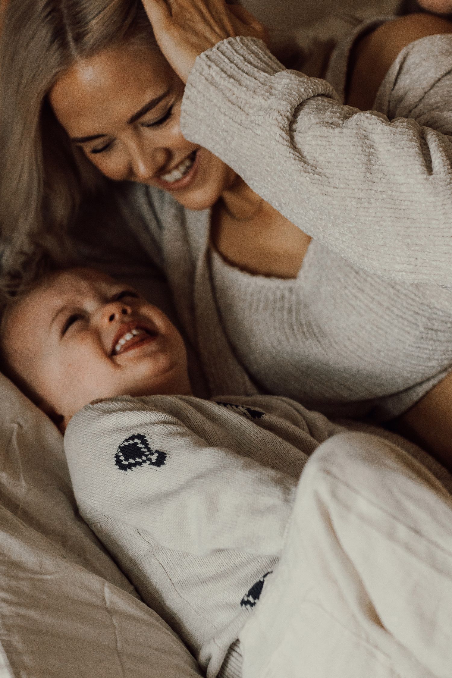 A smiling mother gazes lovingly at her laughing baby lying on a bed, both wearing cozy neutral sweaters.