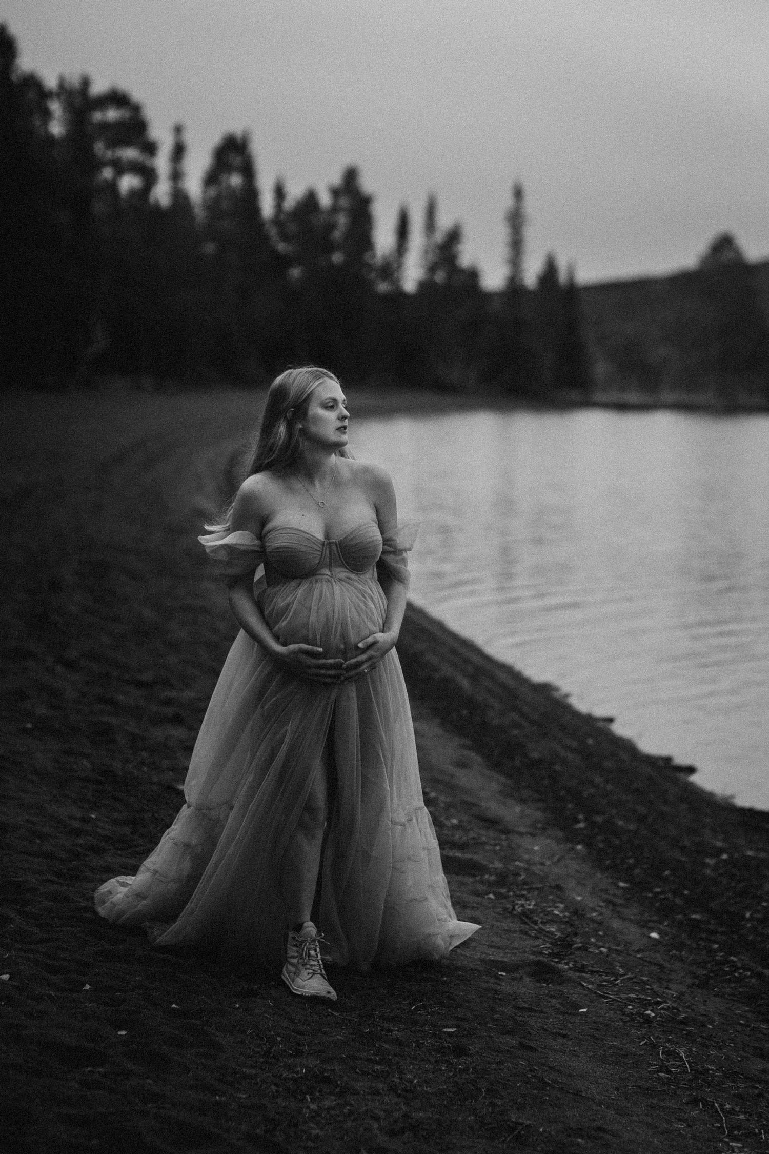 Pregnant woman in elegant off-shoulder tulle gown cradling baby bump by a serene lake, black and white maternity photo.