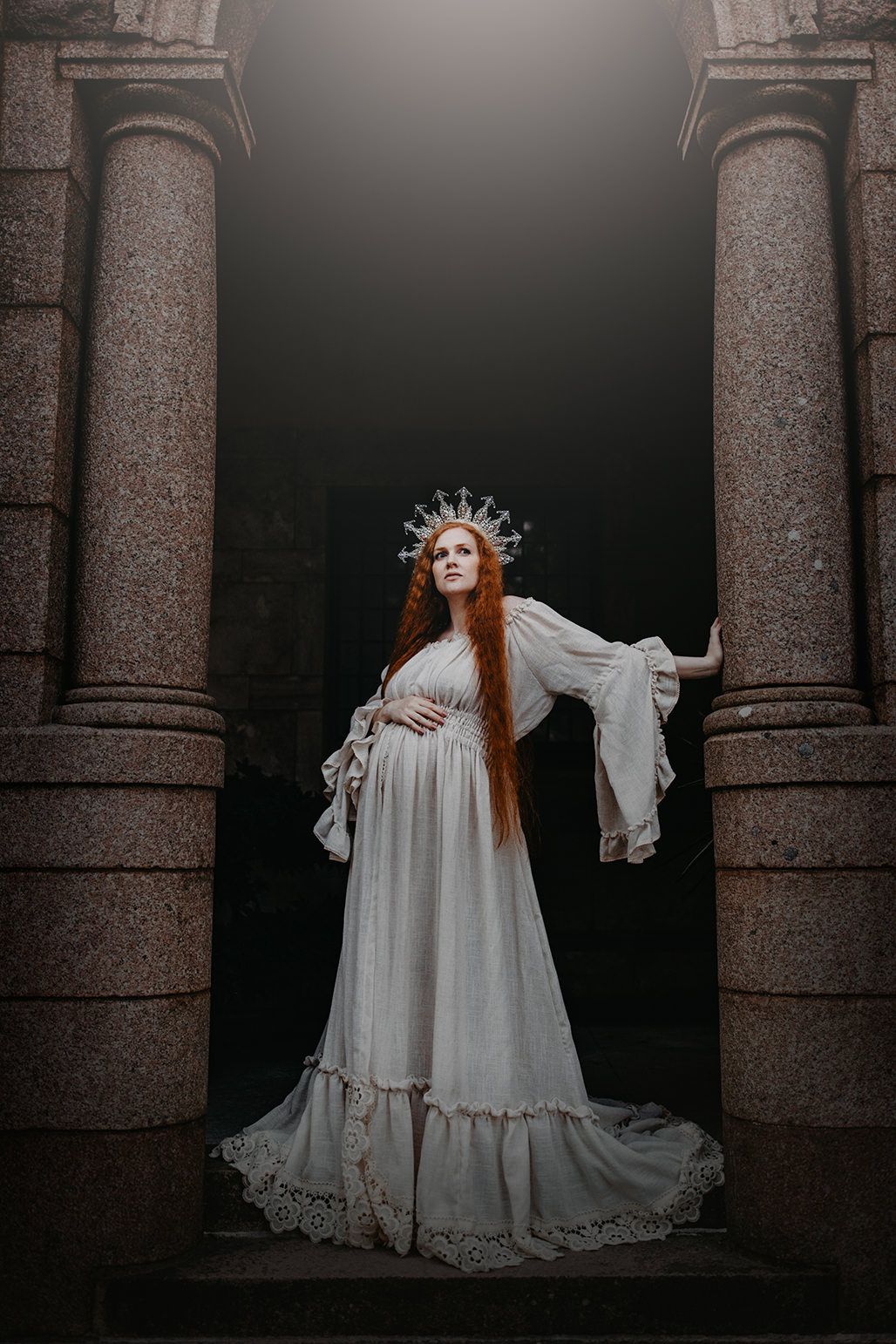 A figure in a flowing white vintage dress and crown poses dramatically between stone columns in a dark gothic setting.