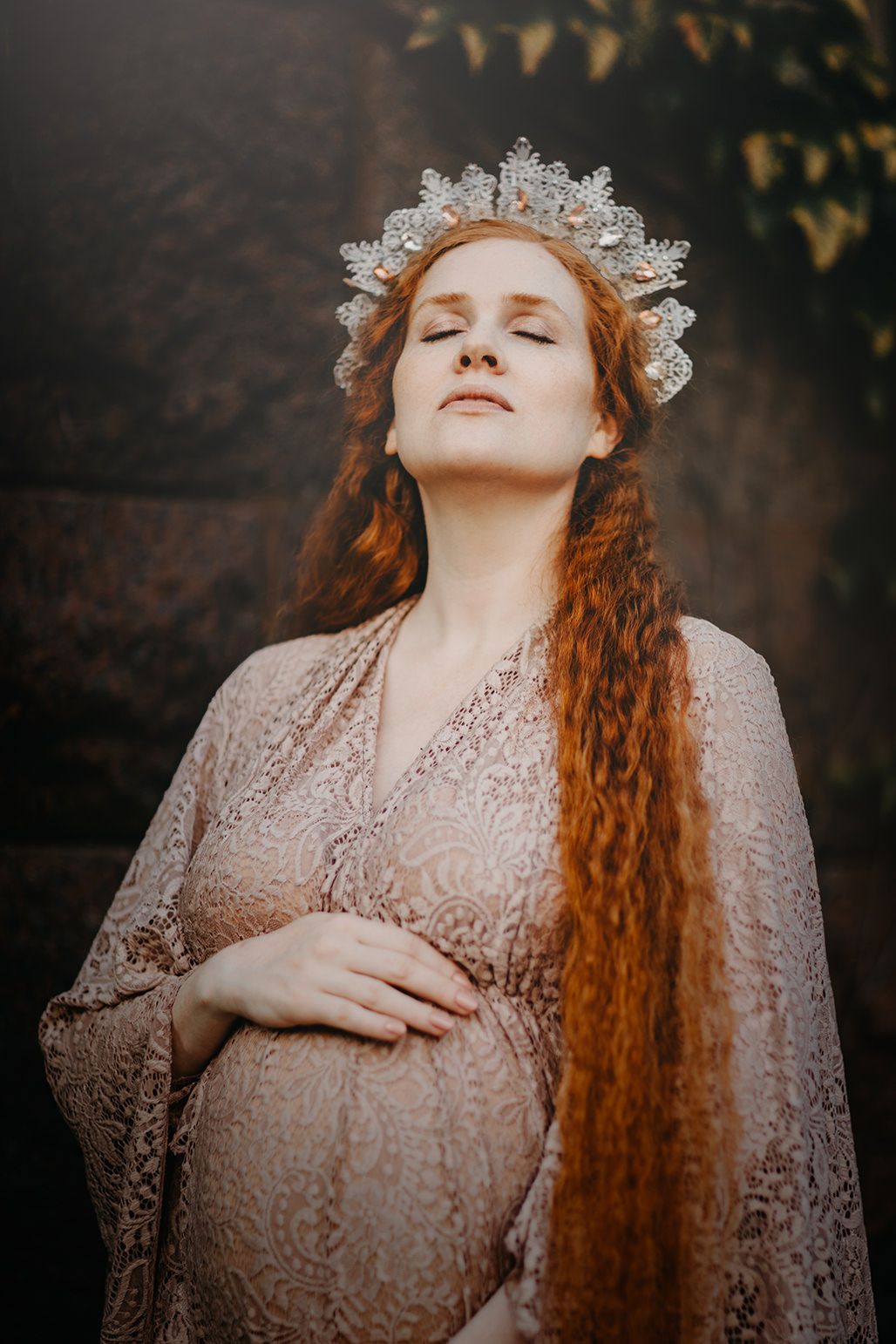 Ethereal maternity portrait with flowing red hair and delicate lace crown in moody outdoor setting.