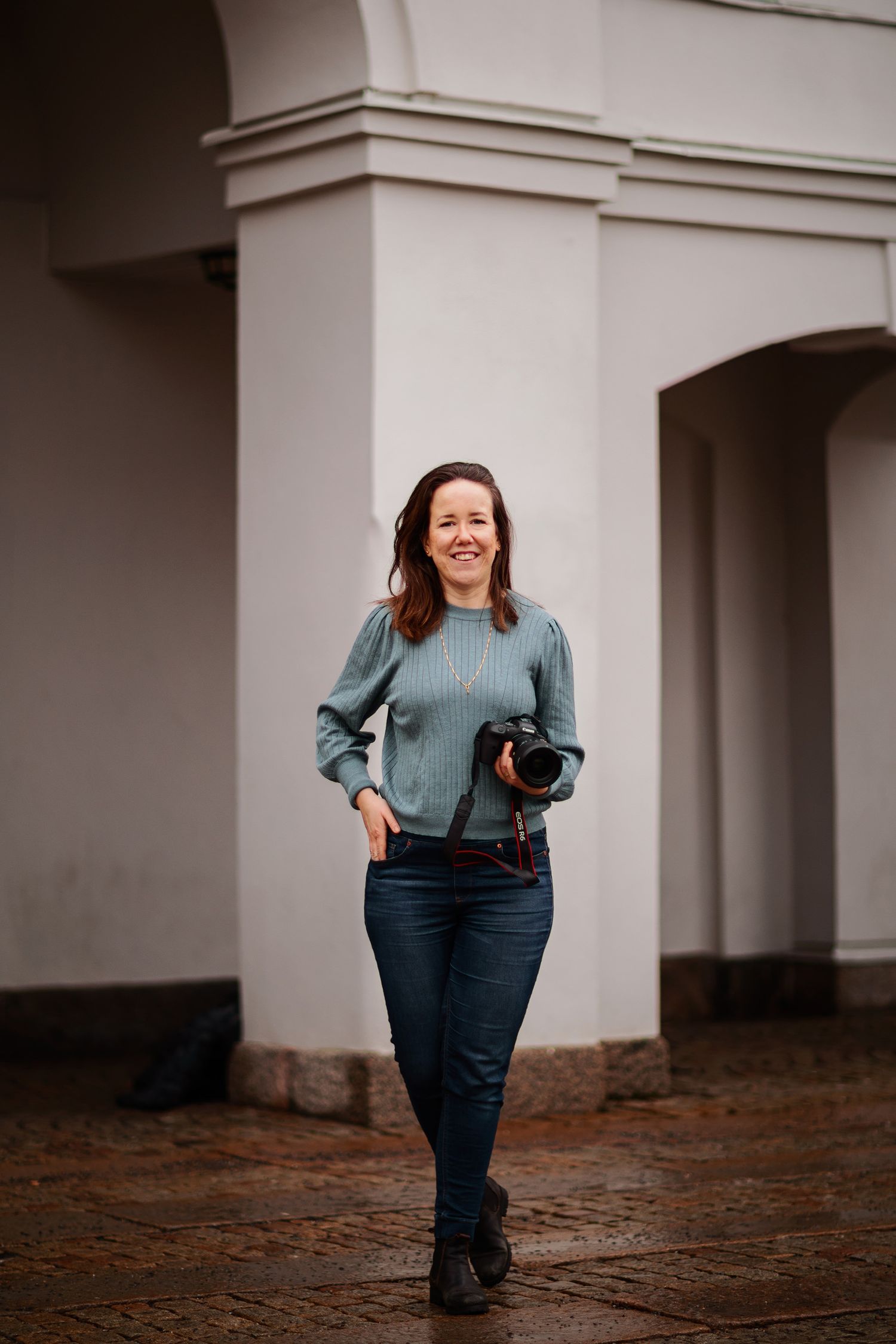 A person wearing a blue sweater and jeans poses against a white architectural column in an outdoor photoshoot.