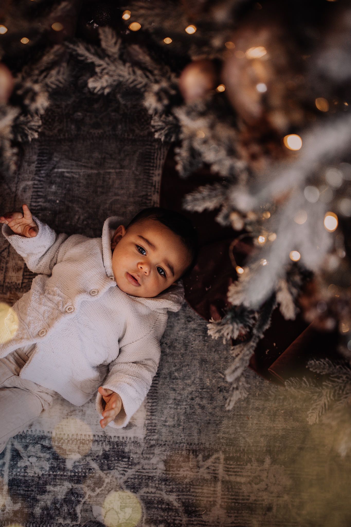 A small child lies underneath sparkling holiday lights and white Christmas tree branches against a dark background.