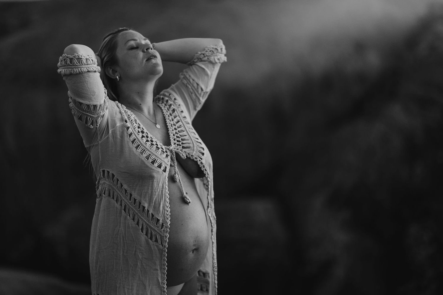 Black and white photo of a pregnant woman in lace top, posing outdoors with eyes closed.