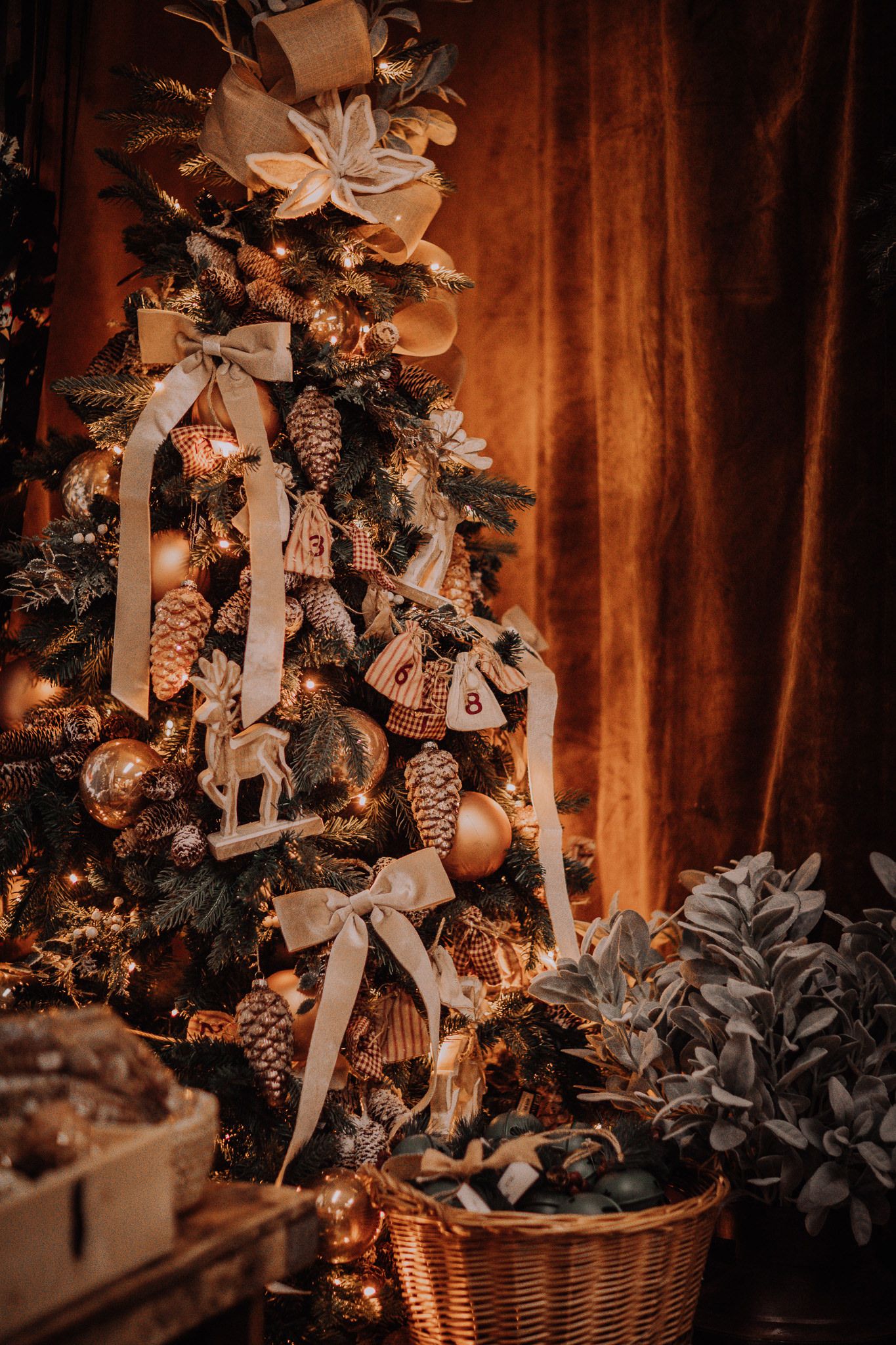 Elegantly decorated Christmas tree with gold ornaments, pine cones, ribbons, and warm lights against rich brown curtains.
