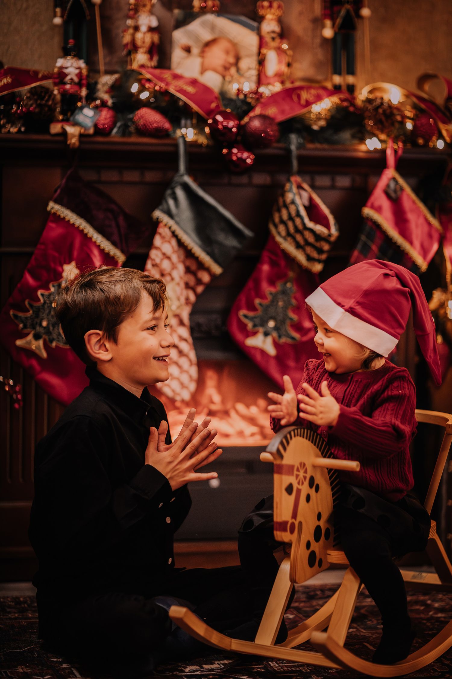 Two children playing together by a decorated Christmas fireplace with stockings, one wearing a Santa hat.