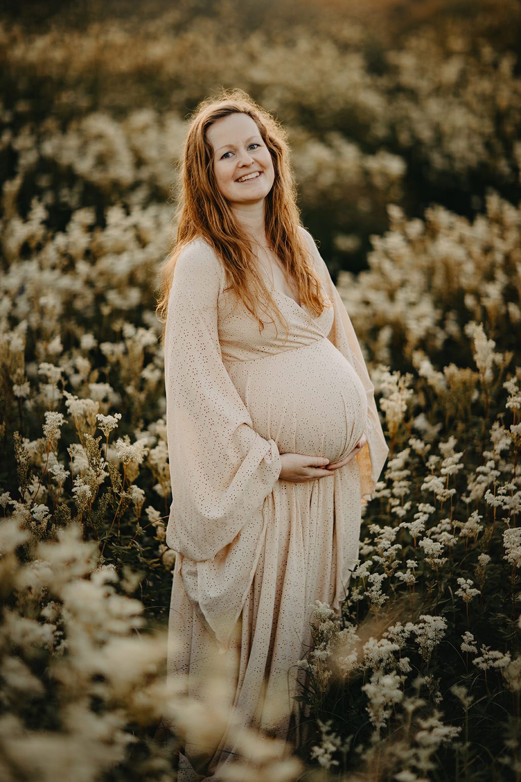 A pregnant woman in a flowy cream dress stands among yellow wildflowers during golden hour.