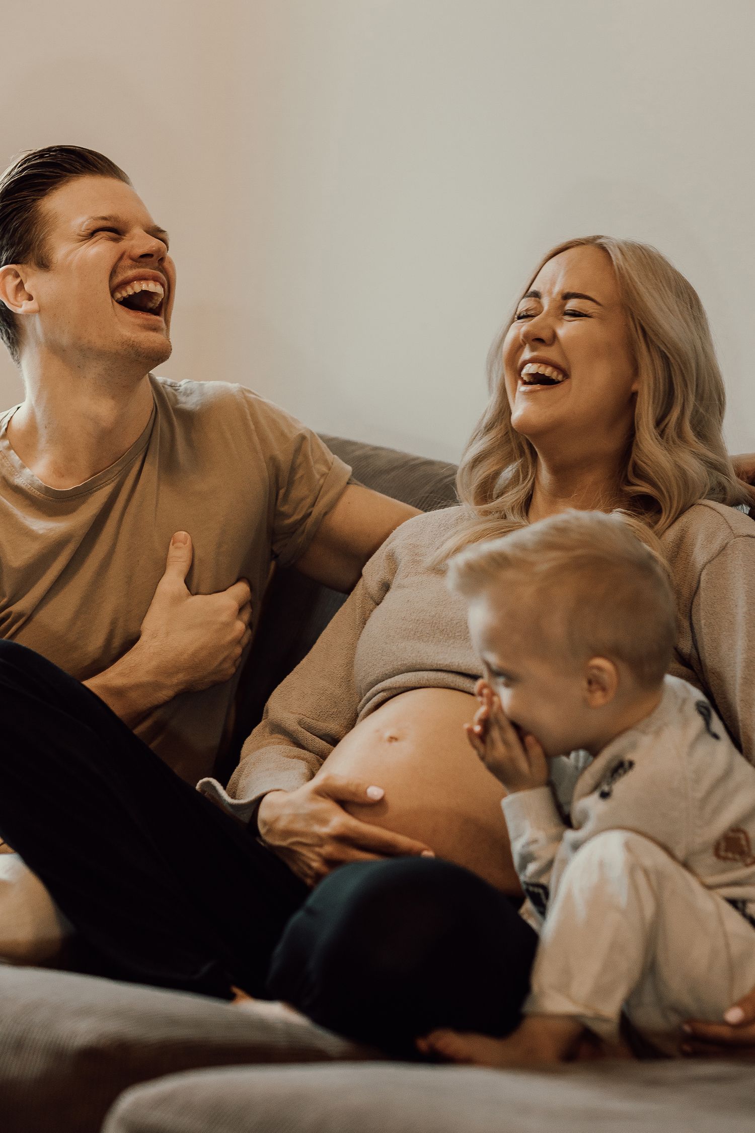 Happy family laughing on couch, pregnant mother with toddler and father sharing a joyful moment together.