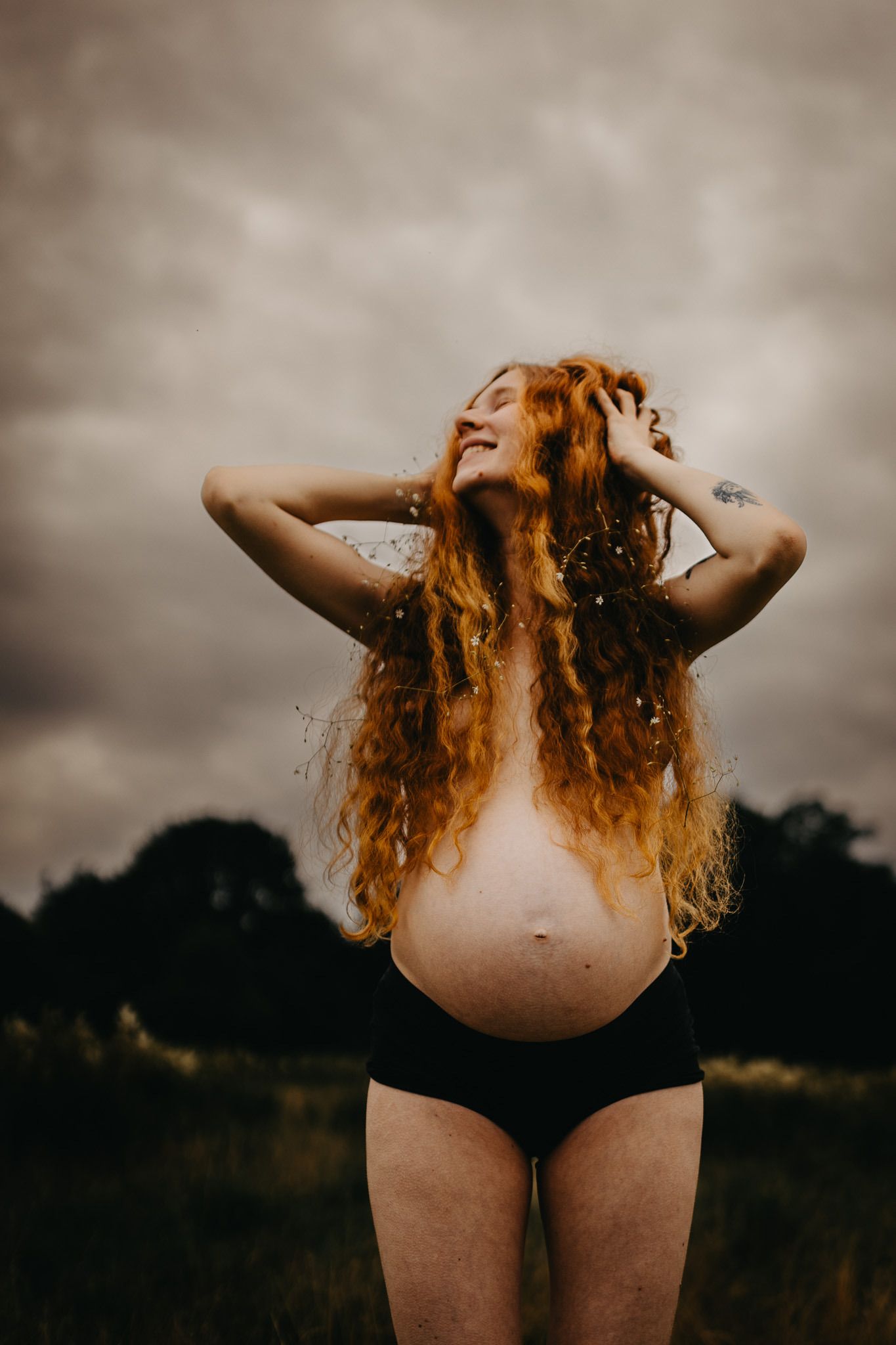 A person with flowing red curly hair flips their head back dramatically against a moody outdoor landscape at sunset.