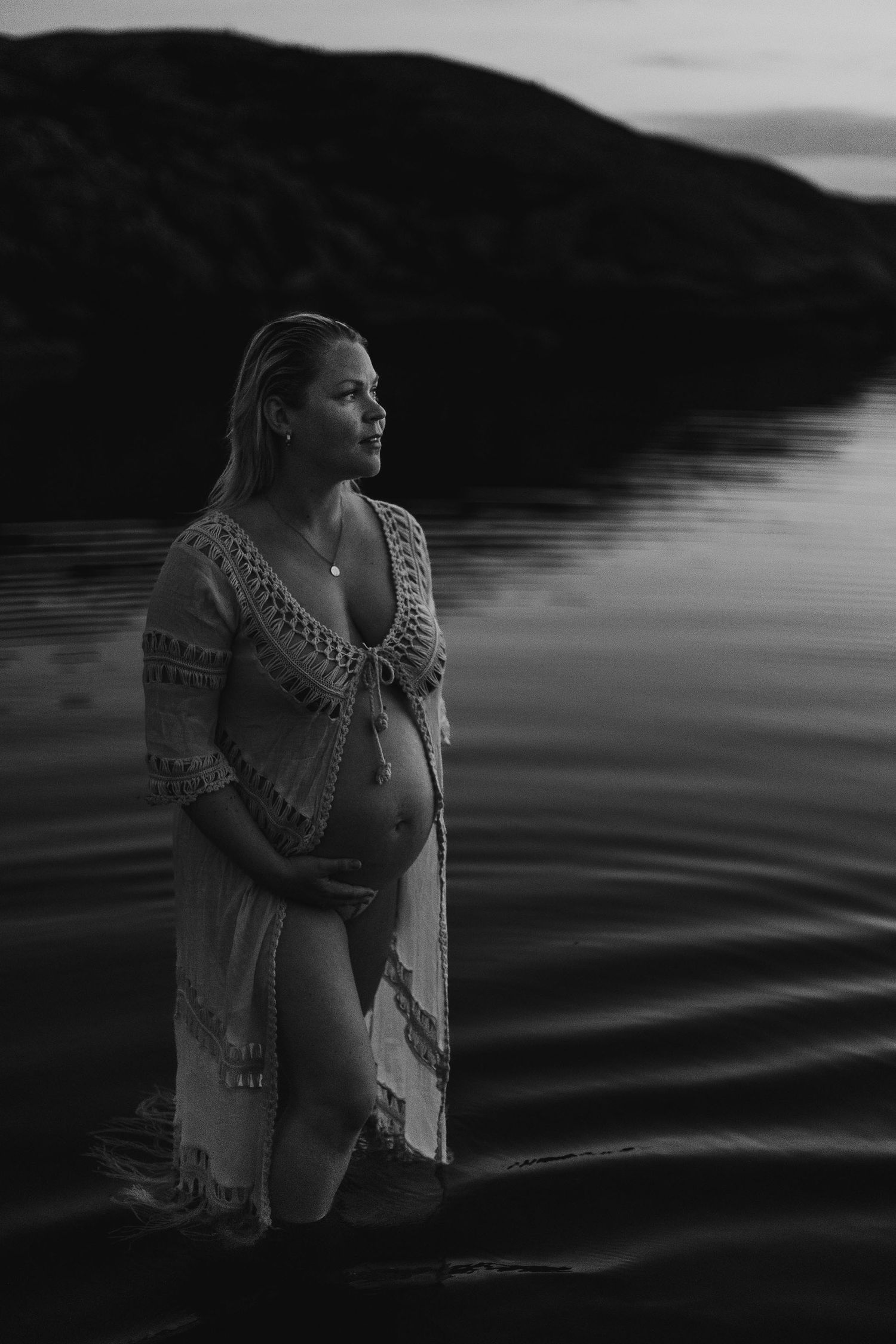 A silhouetted pregnant figure in a lace coverup stands in calm lake waters at dusk with mountains in the background.