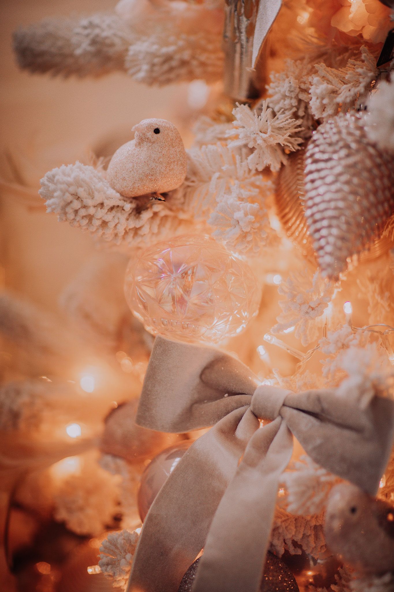 White Christmas tree decorated with glittery bird, pine cones, glass ornament, and silver bow with warm fairy lights.