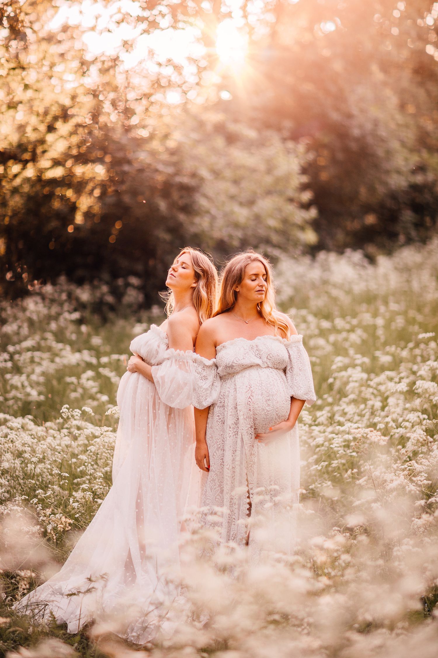 Two women in white dresses standing back-to-back in dreamy meadow of white flowers.
