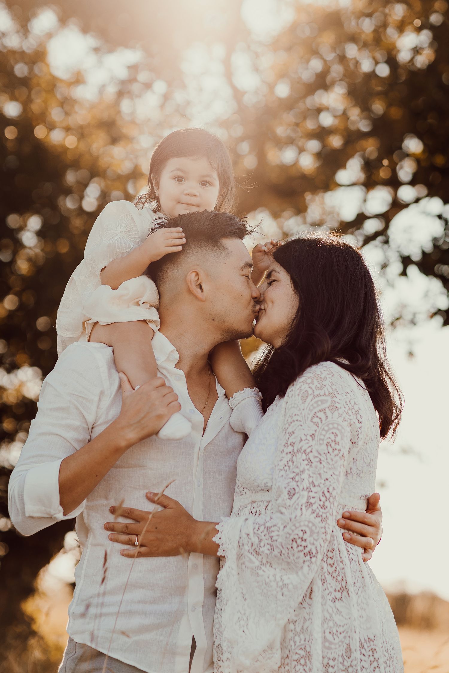 A couple shares a kiss outdoors at golden hour while their toddler sits on dad's shoulders in matching white outfits.