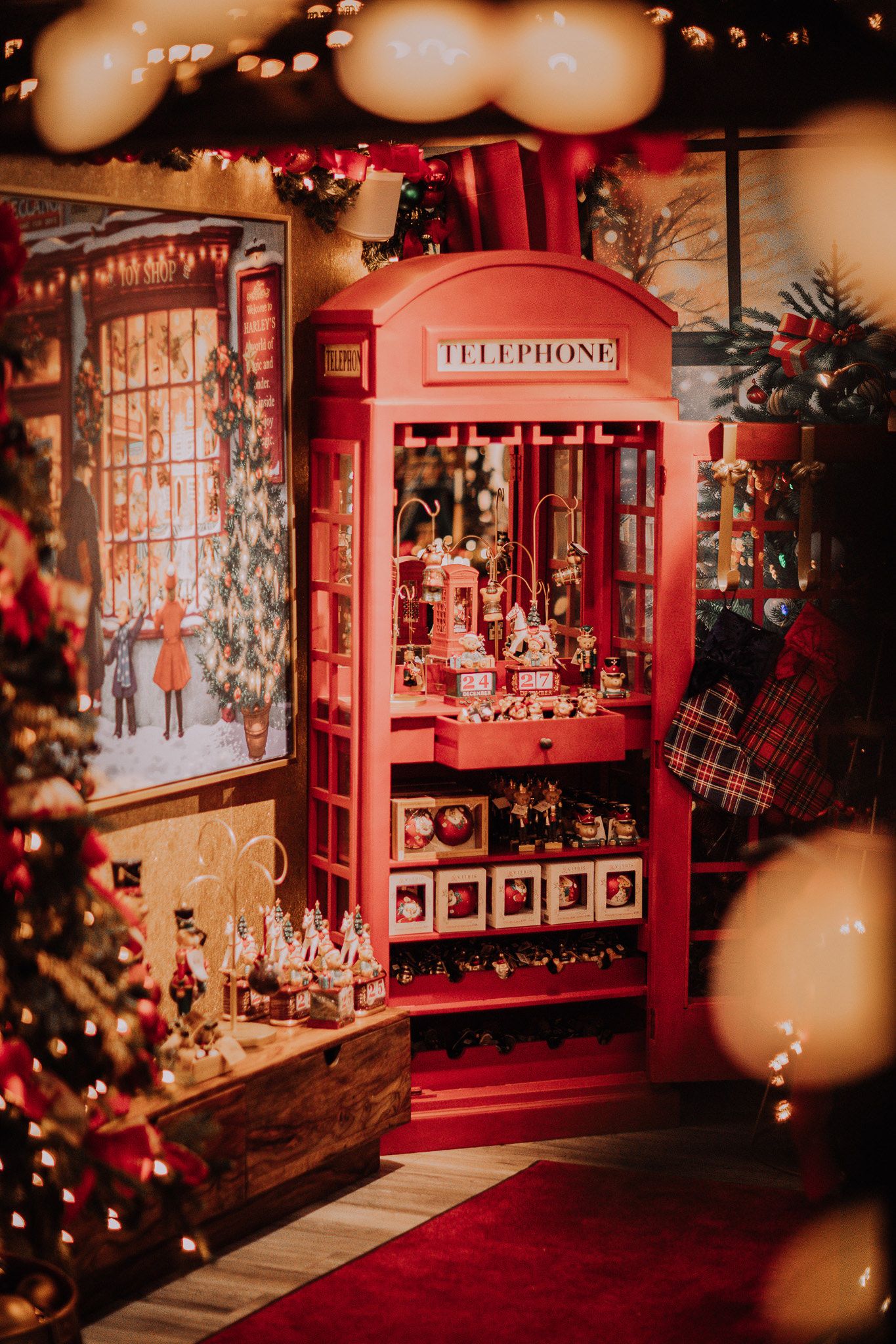 Red British telephone box decorated as a Christmas market stall with festive ornaments, lights, and holiday decor.