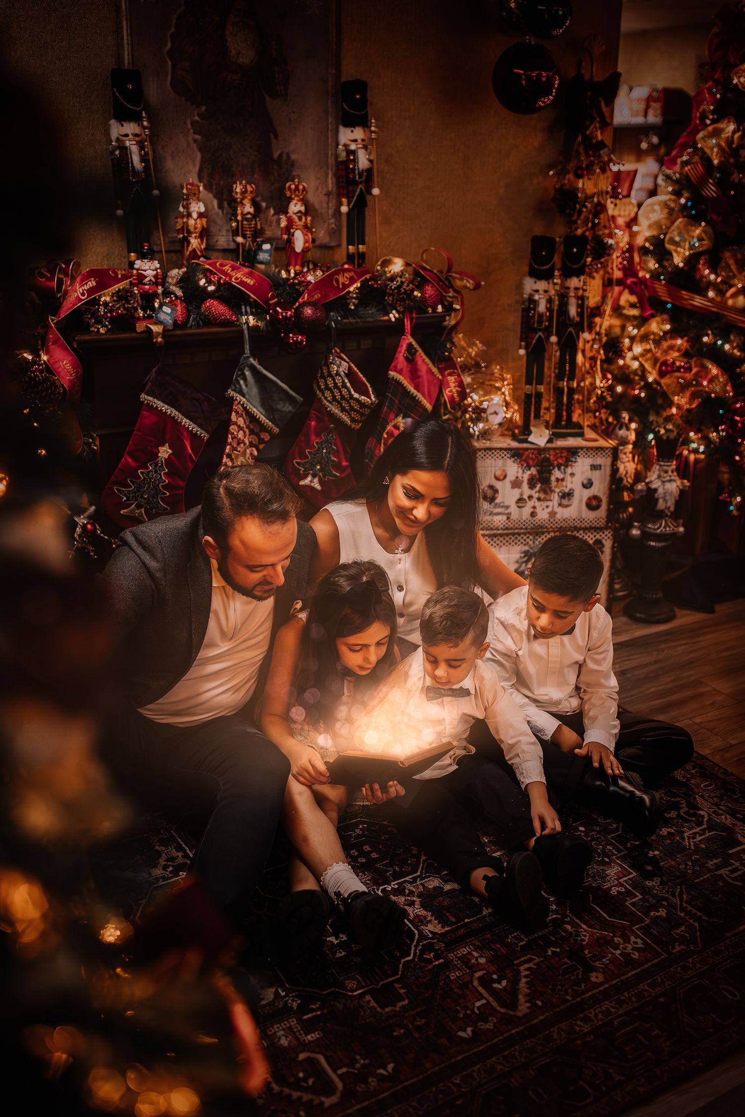Family gathered around lit Christmas tree looking at glowing lights with stockings hung on fireplace mantel in background.