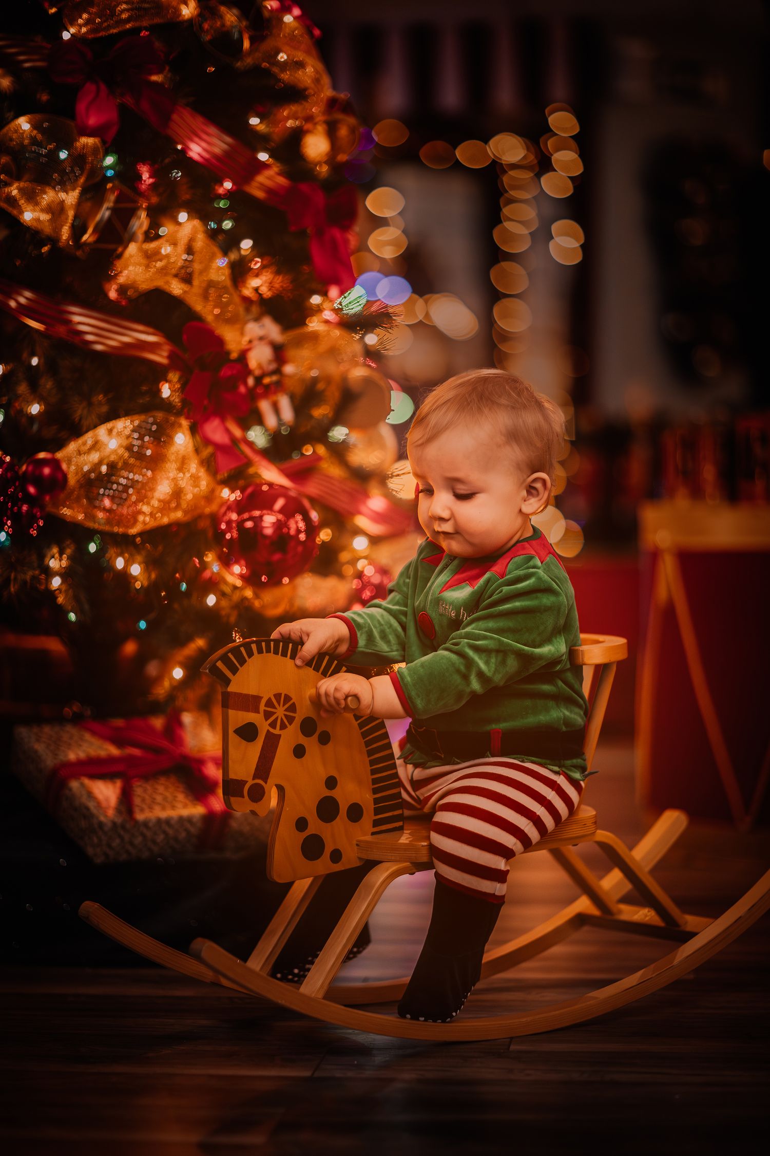 Baby sits in wooden rocking horse near Christmas tree with warm glowing lights in dark room.