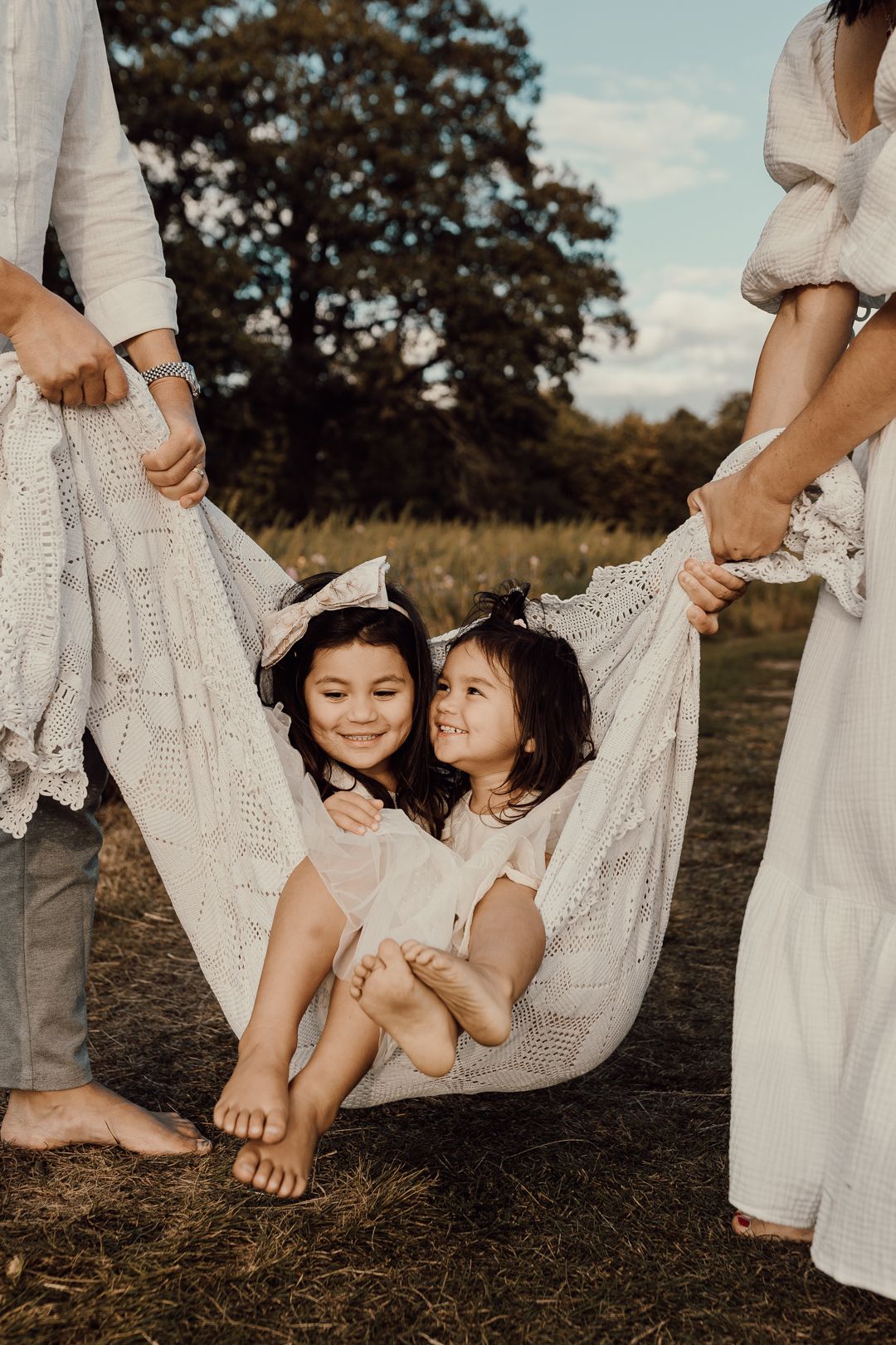 Two young girls in white dresses sit in a blanket hammock held by parents outdoors in a sunny field.