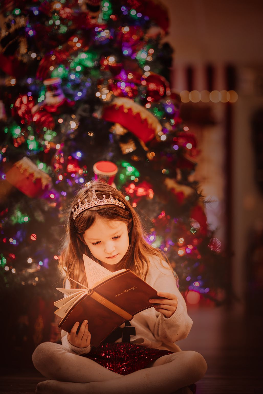 Young girl wearing a crown reads a glowing book while sitting before a decorated Christmas tree with colorful lights.
