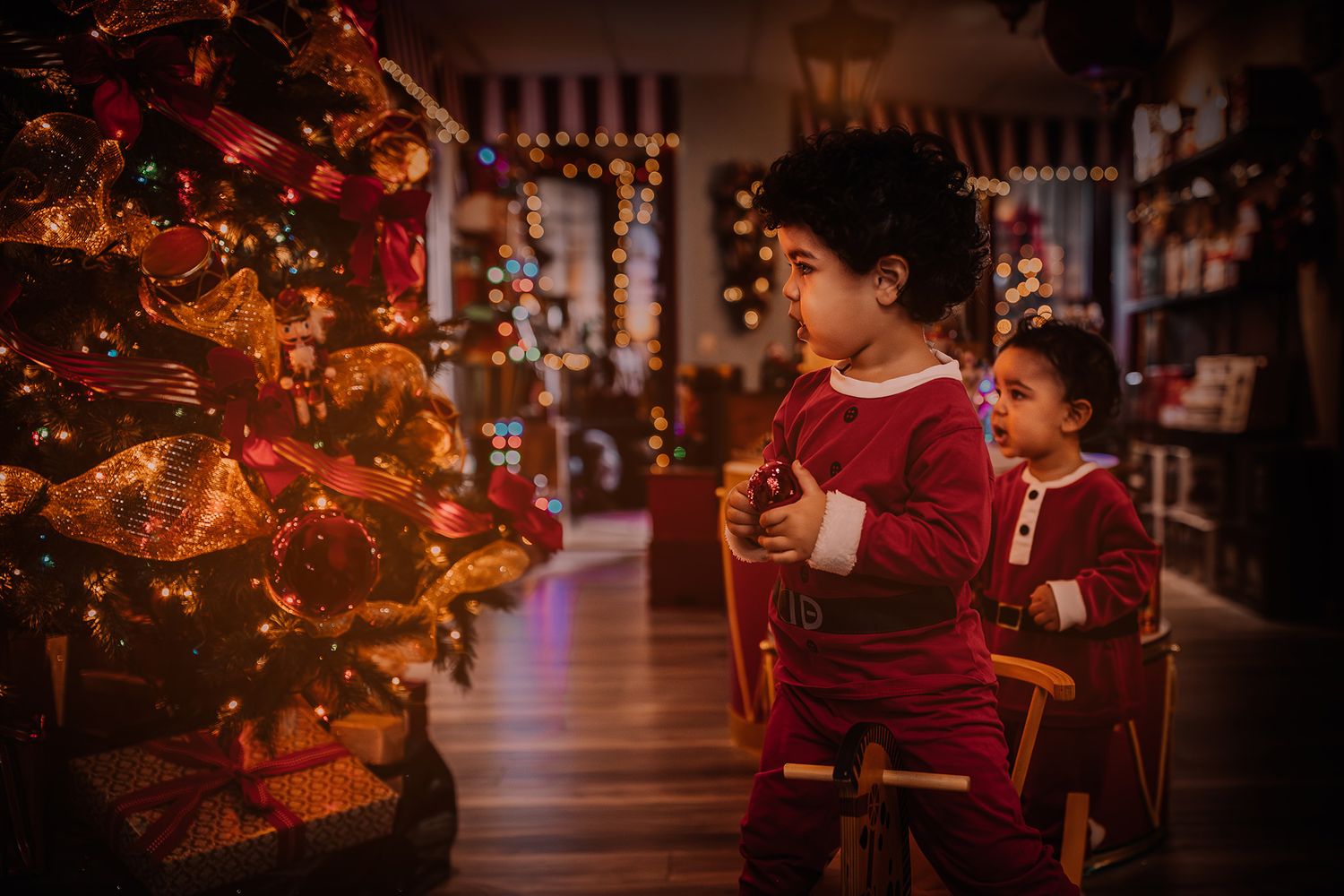 Two young children in red Christmas outfits admire a decorated holiday tree in a warmly lit festive shop.