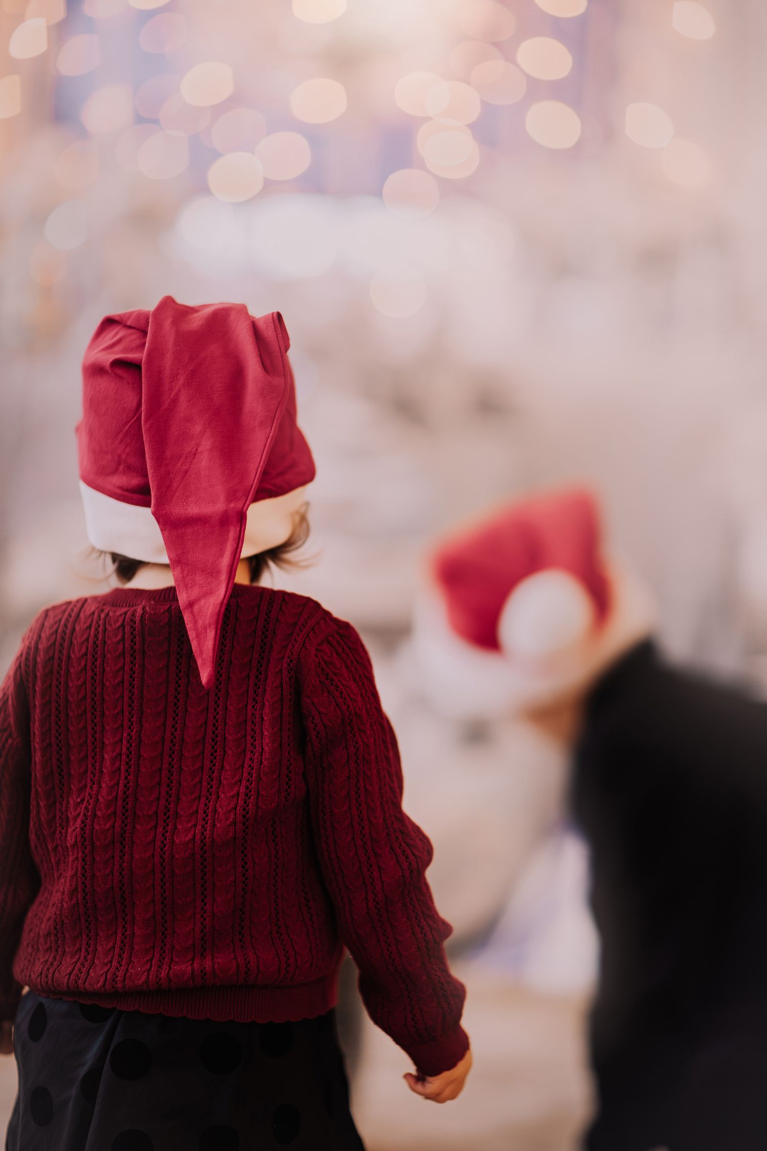 A child in a burgundy sweater and Santa hat stands with their back to the camera against a soft bokeh background.