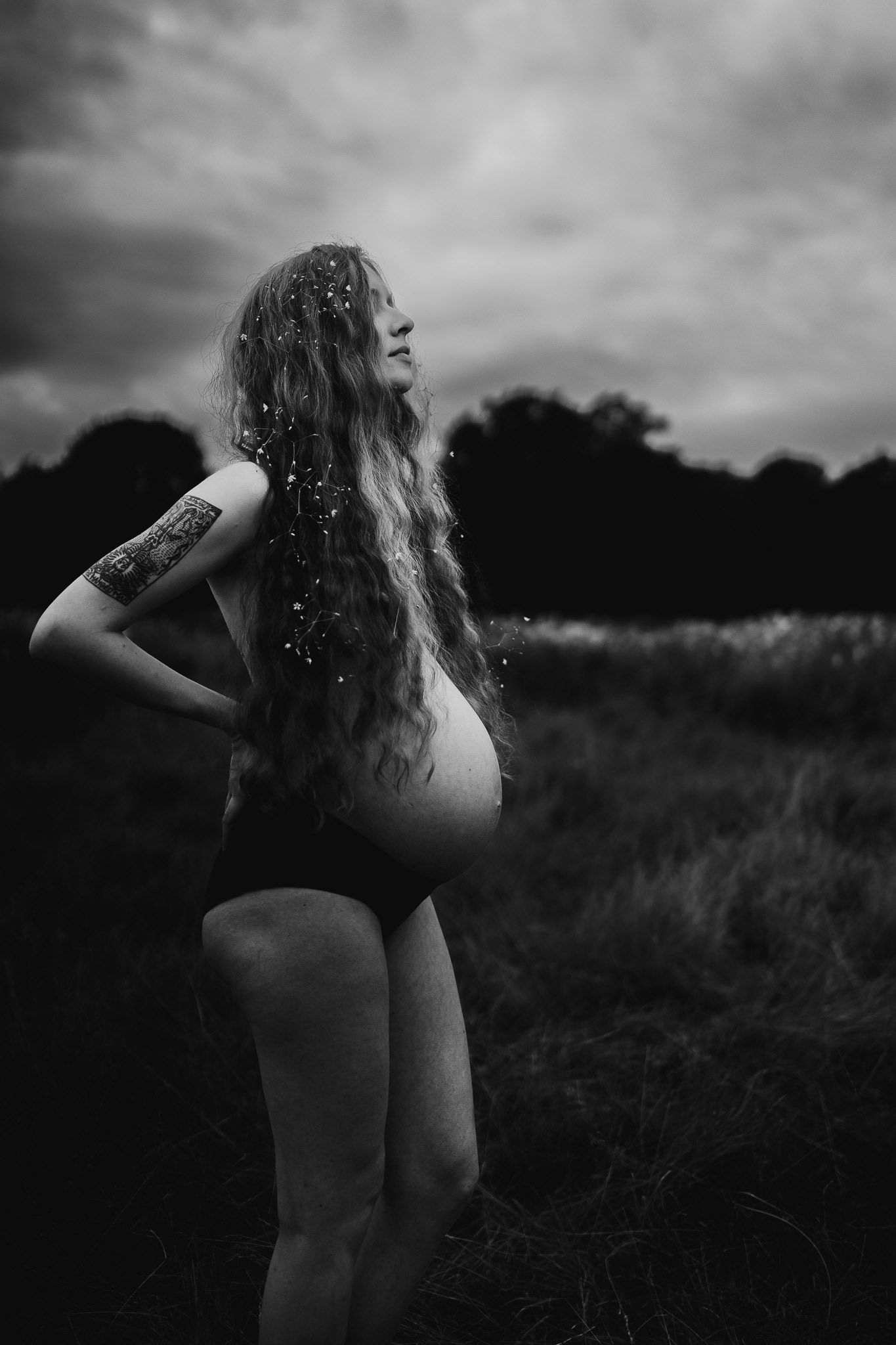 Black and white photo of pregnant woman with long hair and tattoo standing in field under dramatic cloudy sky.