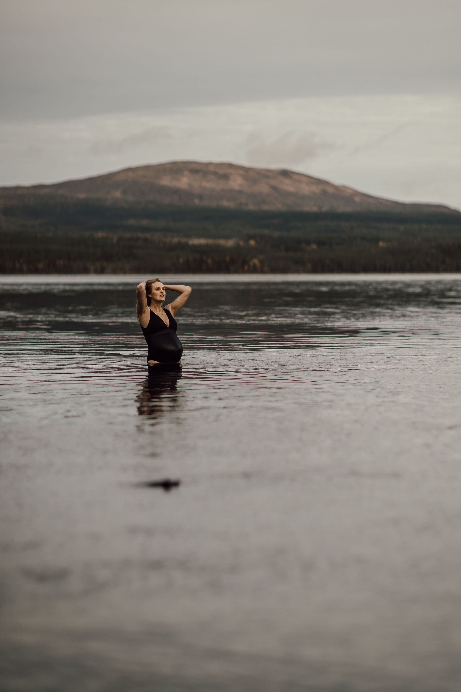 Woman in black swimsuit standing waist-deep in calm lake with mountain and forest backdrop under overcast sky.