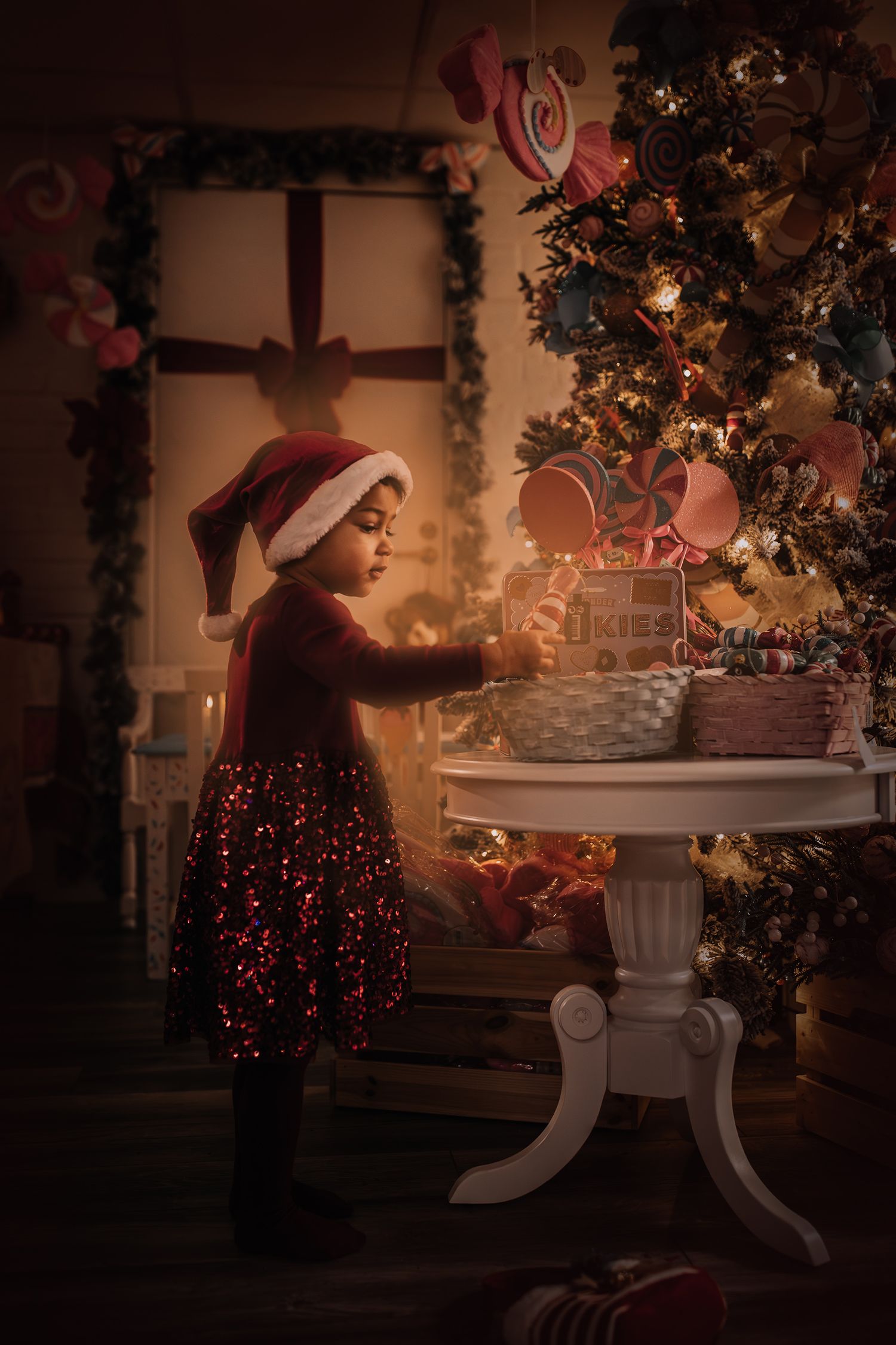 A small child in a Santa hat reaches for decorations on a white table near a Christmas tree.