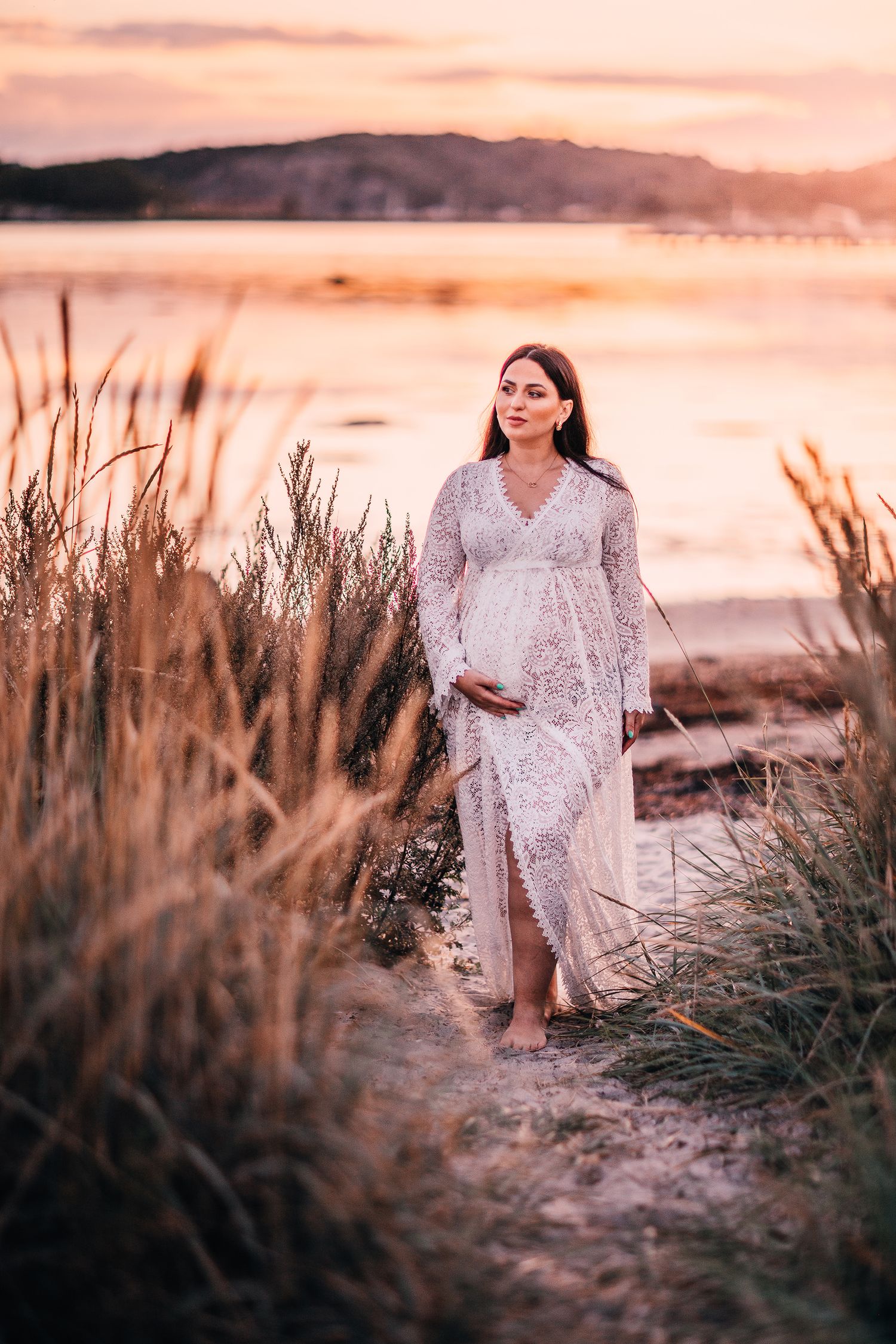 Expectant mother in lace dress walking along lakeside at sunset.