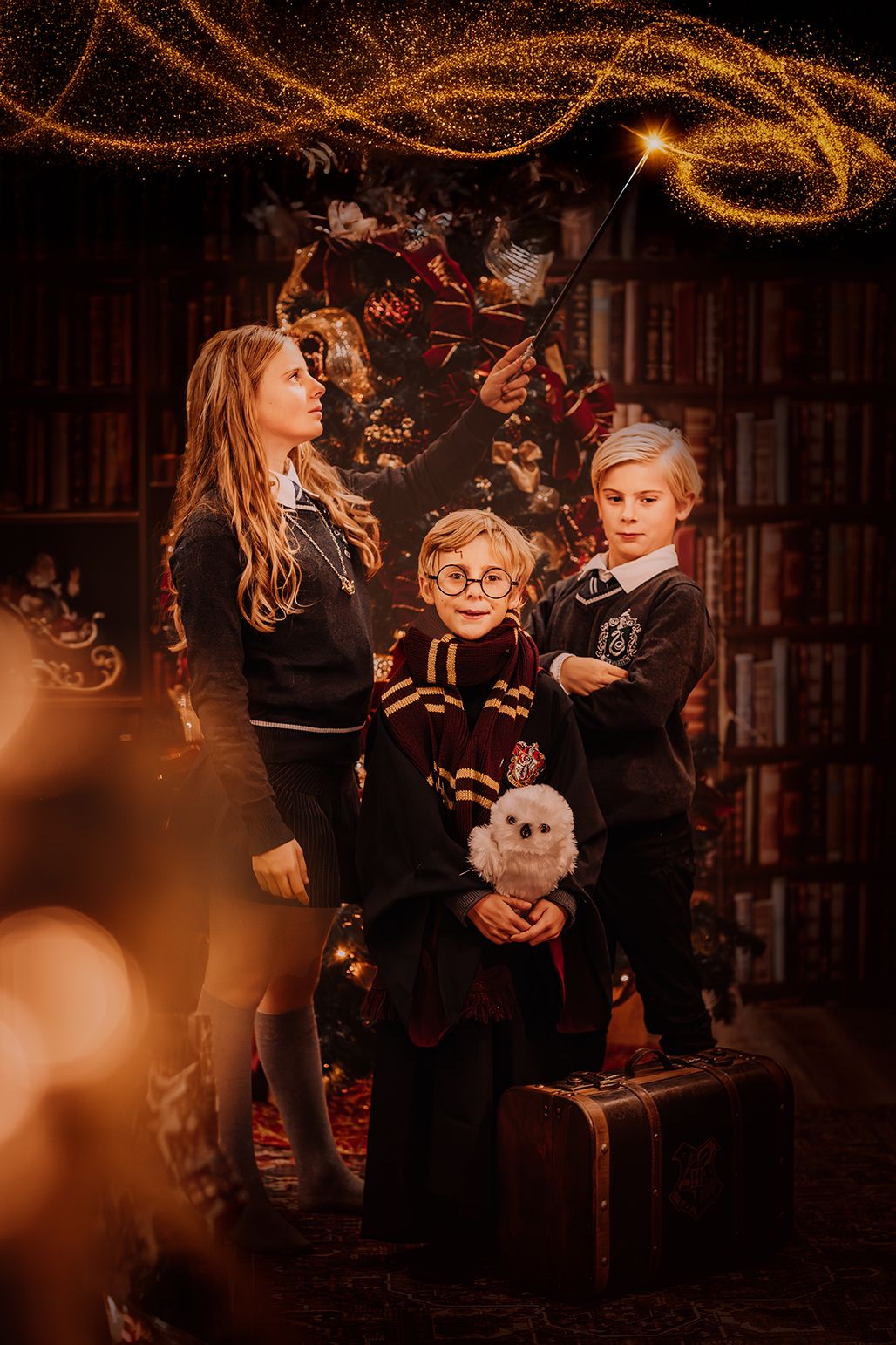 Three children in Harry Potter costumes pose with a wand, owl, and trunk in a magical glowing library setting.