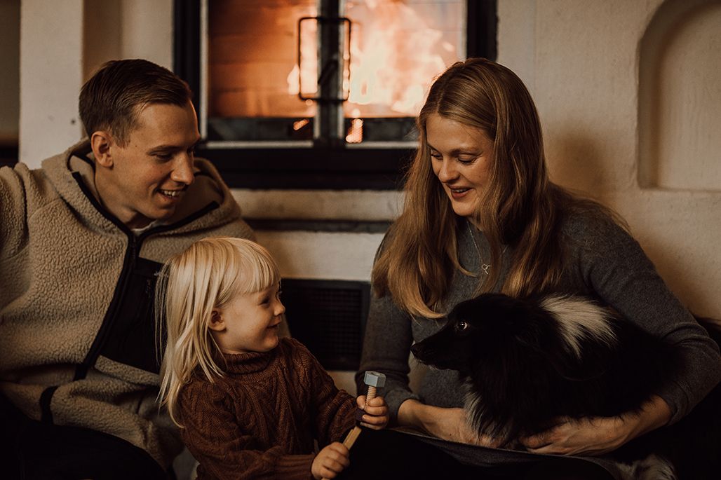 Family with young child and black-white dog sitting by cozy fireplace in warm living room setting.