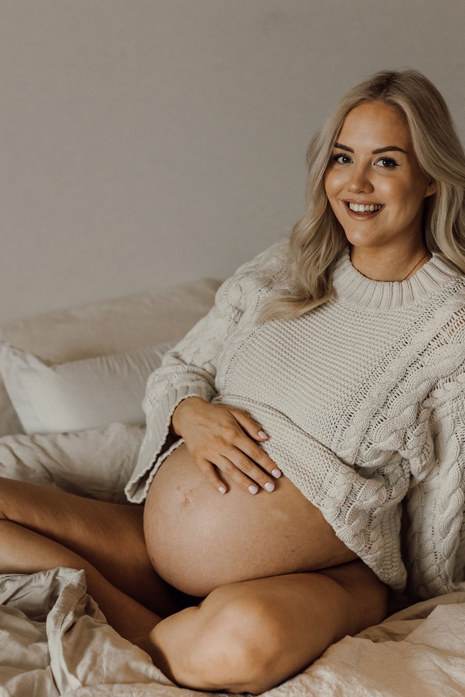 Smiling pregnant woman in cream cable-knit sweater sitting on bed, cradling baby bump with hands.