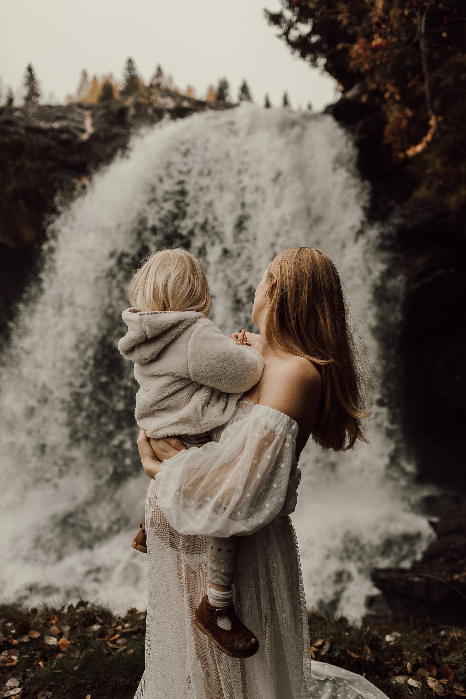 Woman in white dress holding a toddler before a rushing waterfall surrounded by autumn trees.