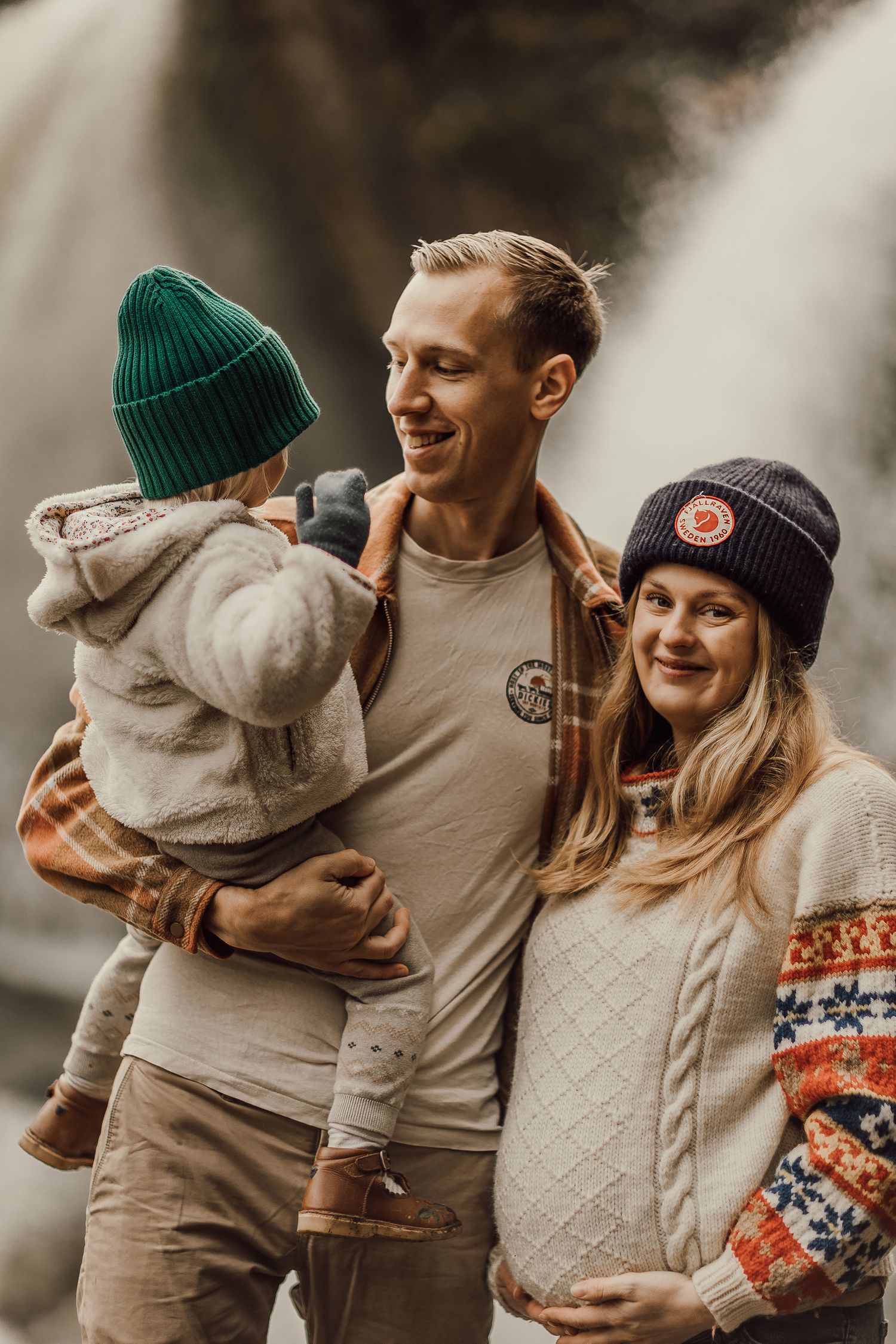 Happy family with pregnant mother, father, and toddler posing outdoors in cozy fall attire near a waterfall.