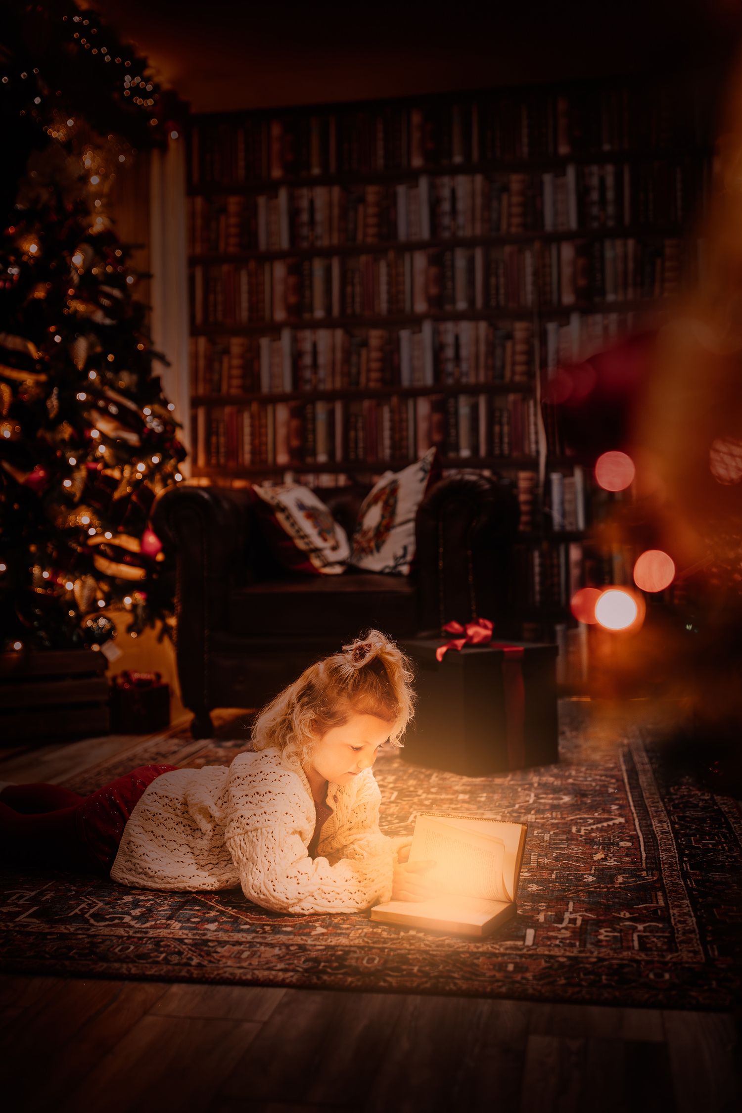 A cozy Christmas scene with someone reading a glowing book on a rug beside a lit tree and bookshelf wall.