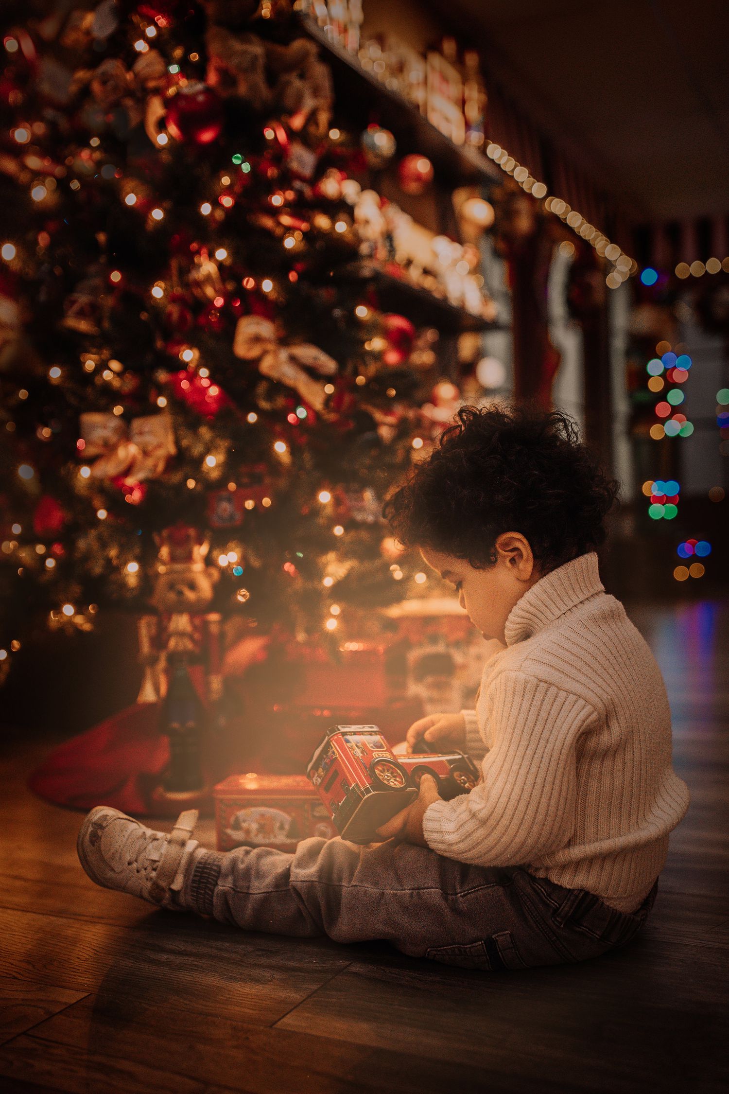 A child in a white sweater sits on the floor examining a gift near a lit Christmas tree.