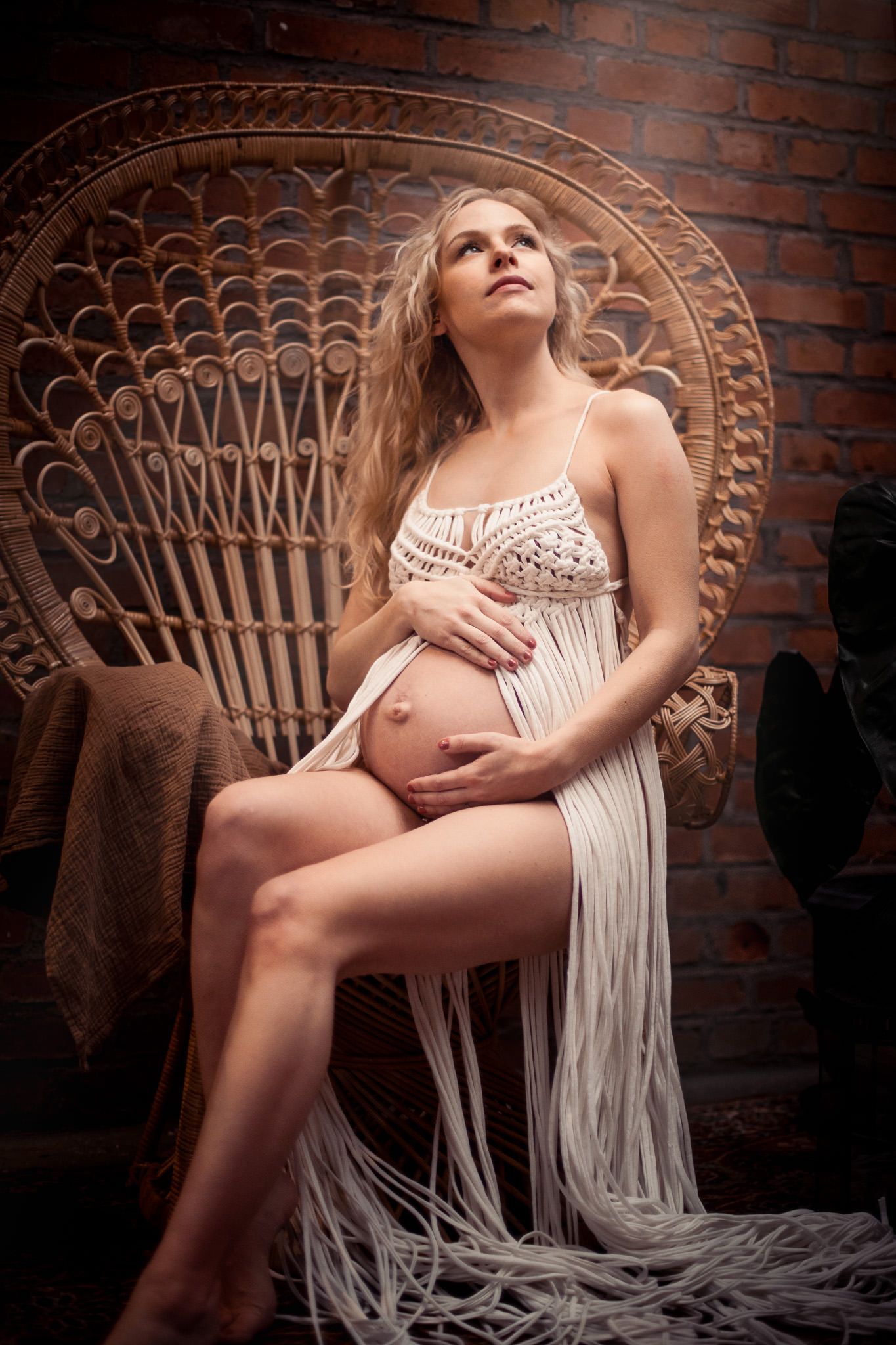 Pregnant woman in white macrame dress sitting in wicker peacock chair against brick wall, cradling baby bump.