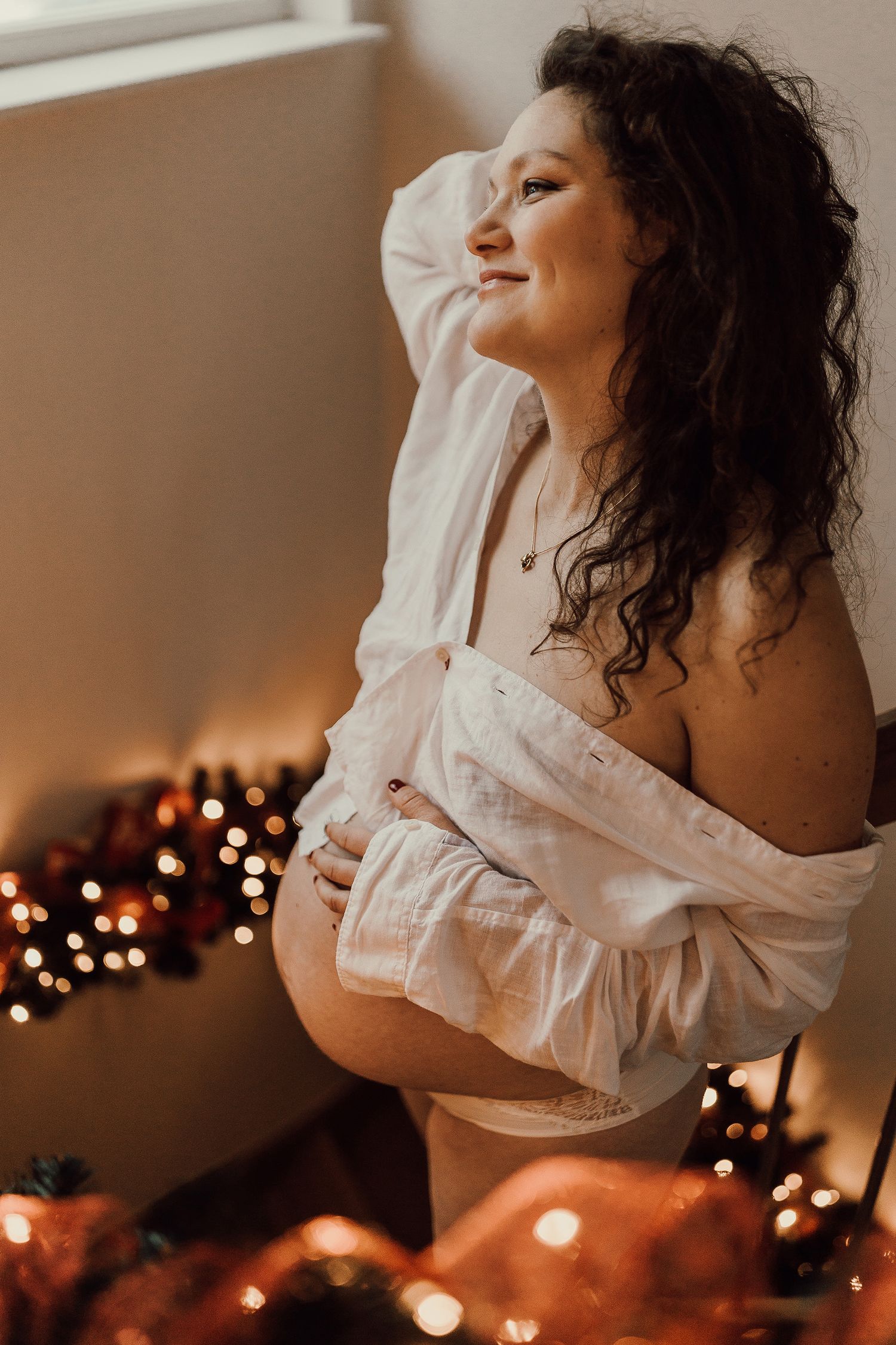 Smiling pregnant woman in white off-shoulder shirt cradling belly, surrounded by warm Christmas lights and ornaments.