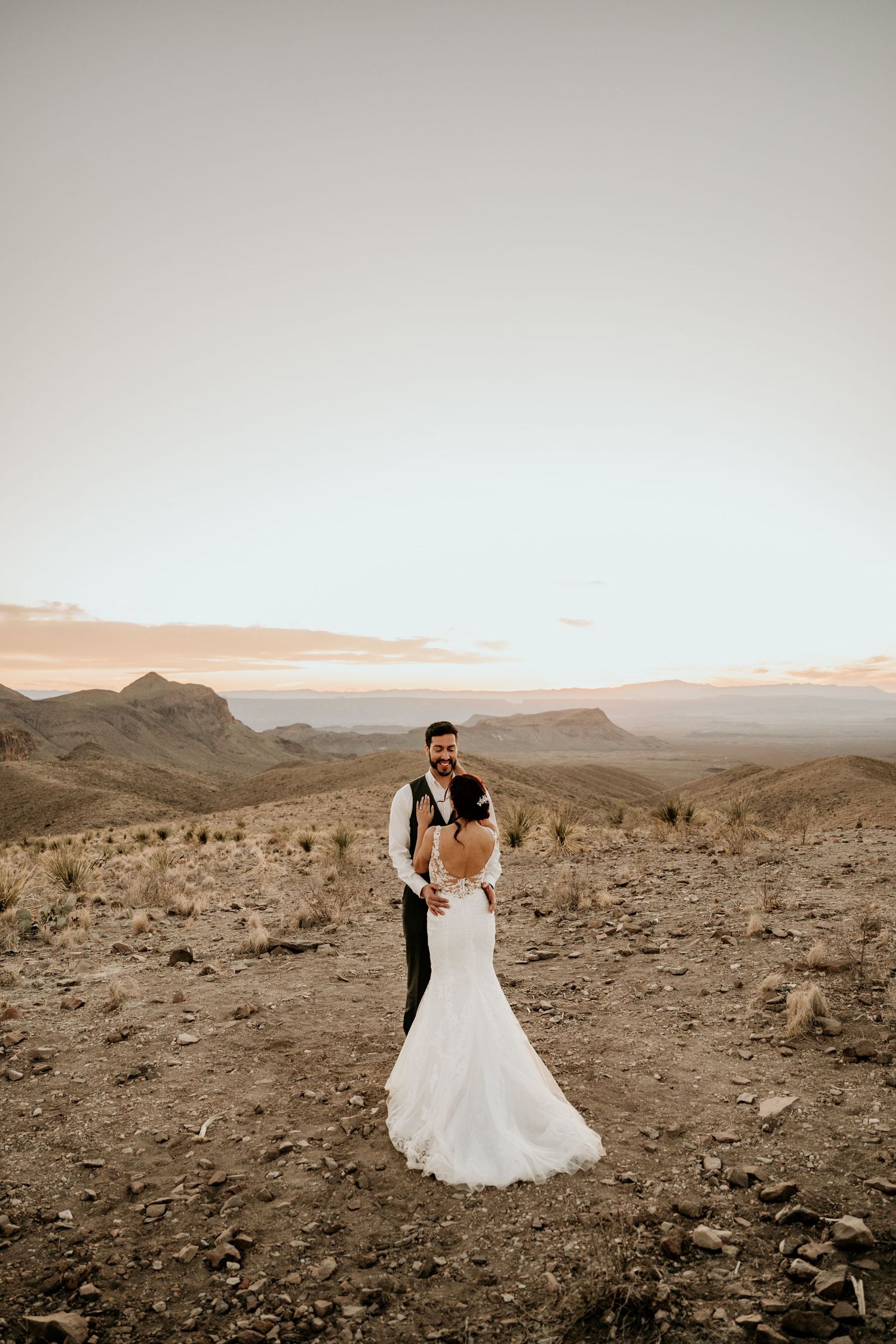 Wedding couple embraces in desert landscape with mountains in background during sunset.